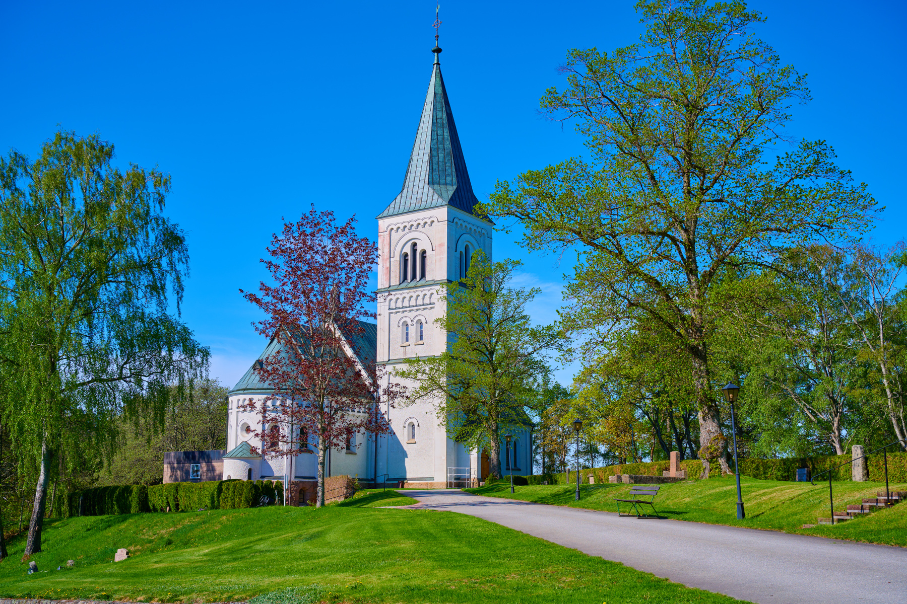 En vit kyrka med ett grått spetsigt tak som reser sig mot en klarblå himmel. Runt kyrkan finns gröna gräsmattor och lövträd med grönskande blad. En asfalterad väg leder upp mot kyrkan, och en bänk syns vid sidan av vägen. En häck löper längs gräsmatt