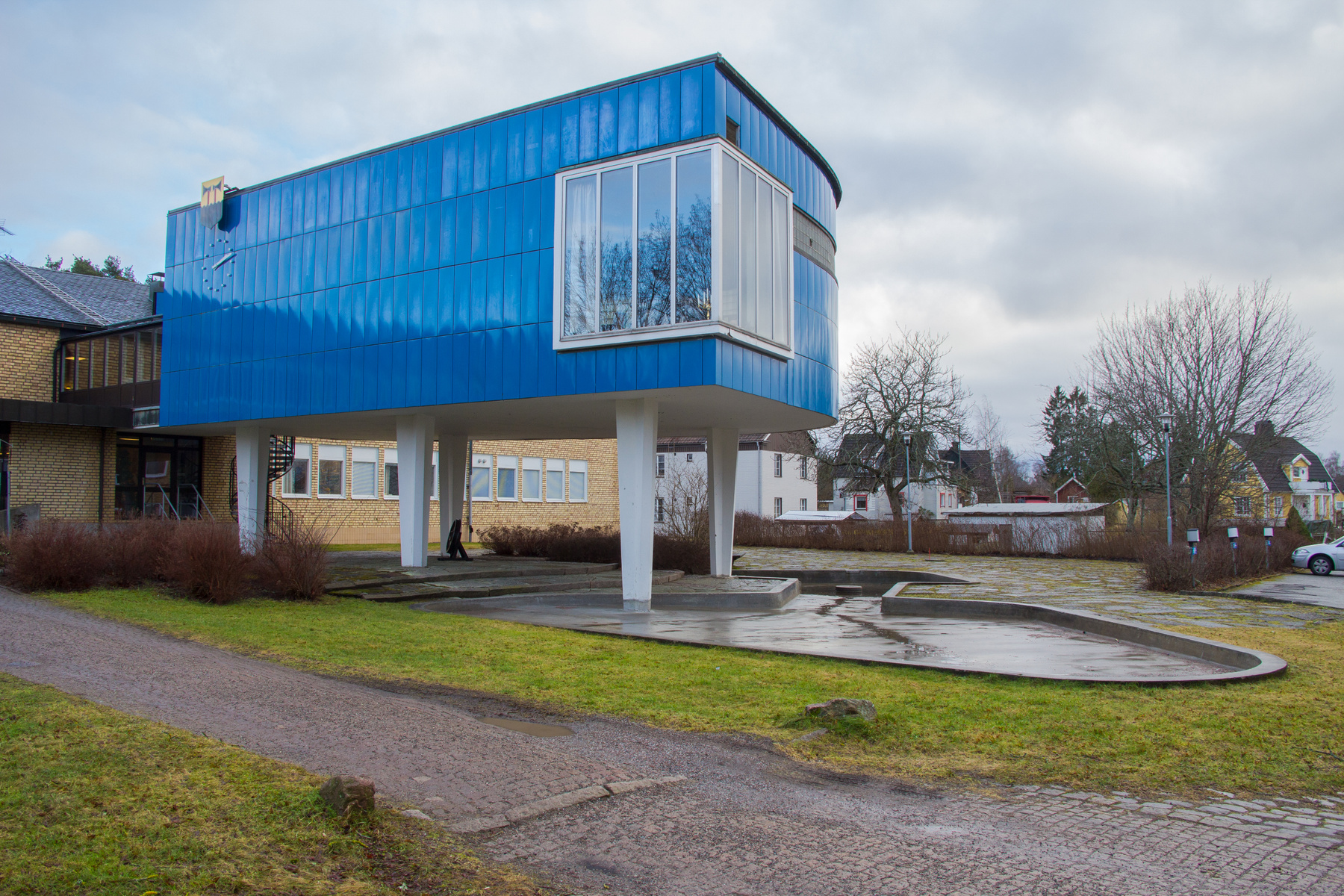 En modern byggnad med en utskjutande övre våning klädd i blå panel, uppburen av vita pelare, står intill en tegelbyggnad under en molnig himmel. Framför finns gräsytor, en stenbelagd gångväg och en betonganläggning. I bakgrunden syns villor och kala