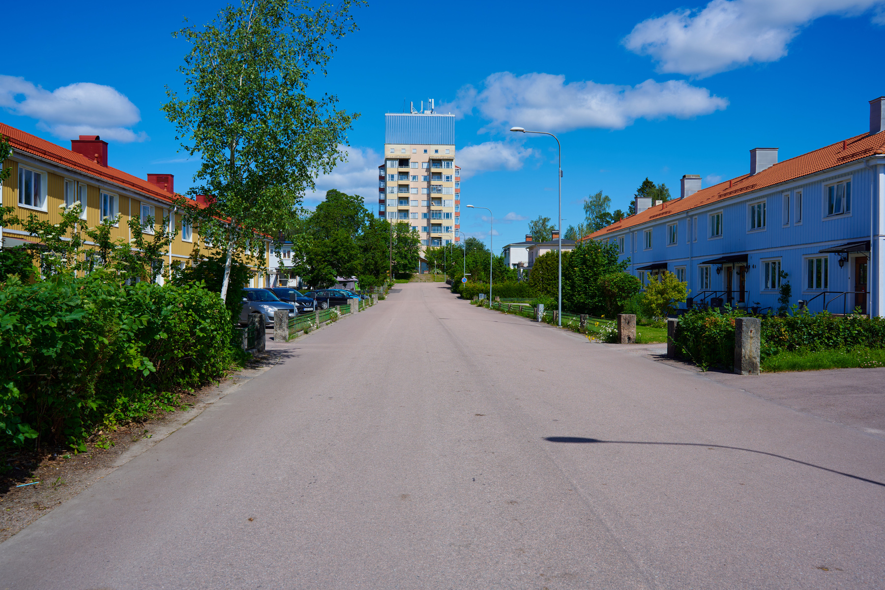 En gata med parkerade bilar och flerfamiljshus på båda sidor, under en klarblå himmel med spridda moln. Längst bort i bild syns ett högre bostadshus. Grönska som buskar och träd kantar gatan och husen.