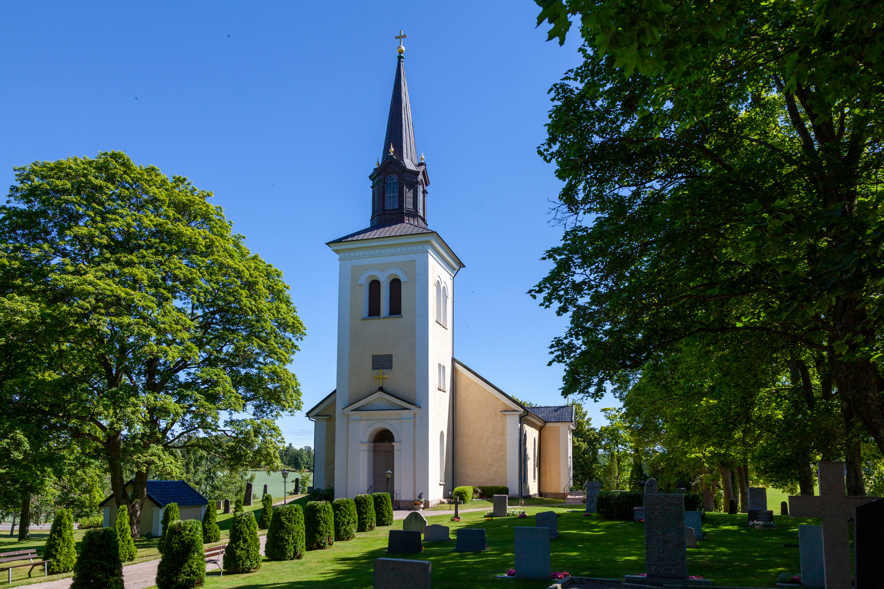 En kyrkobyggnad med ett högt torn och spetsig spira står i centrum av bilden, omgiven av en kyrkogård med gröna gräsmattor och gravstenar. Stora gröna träd ramar in kyrkan på båda sidor under en klarblå himmel.