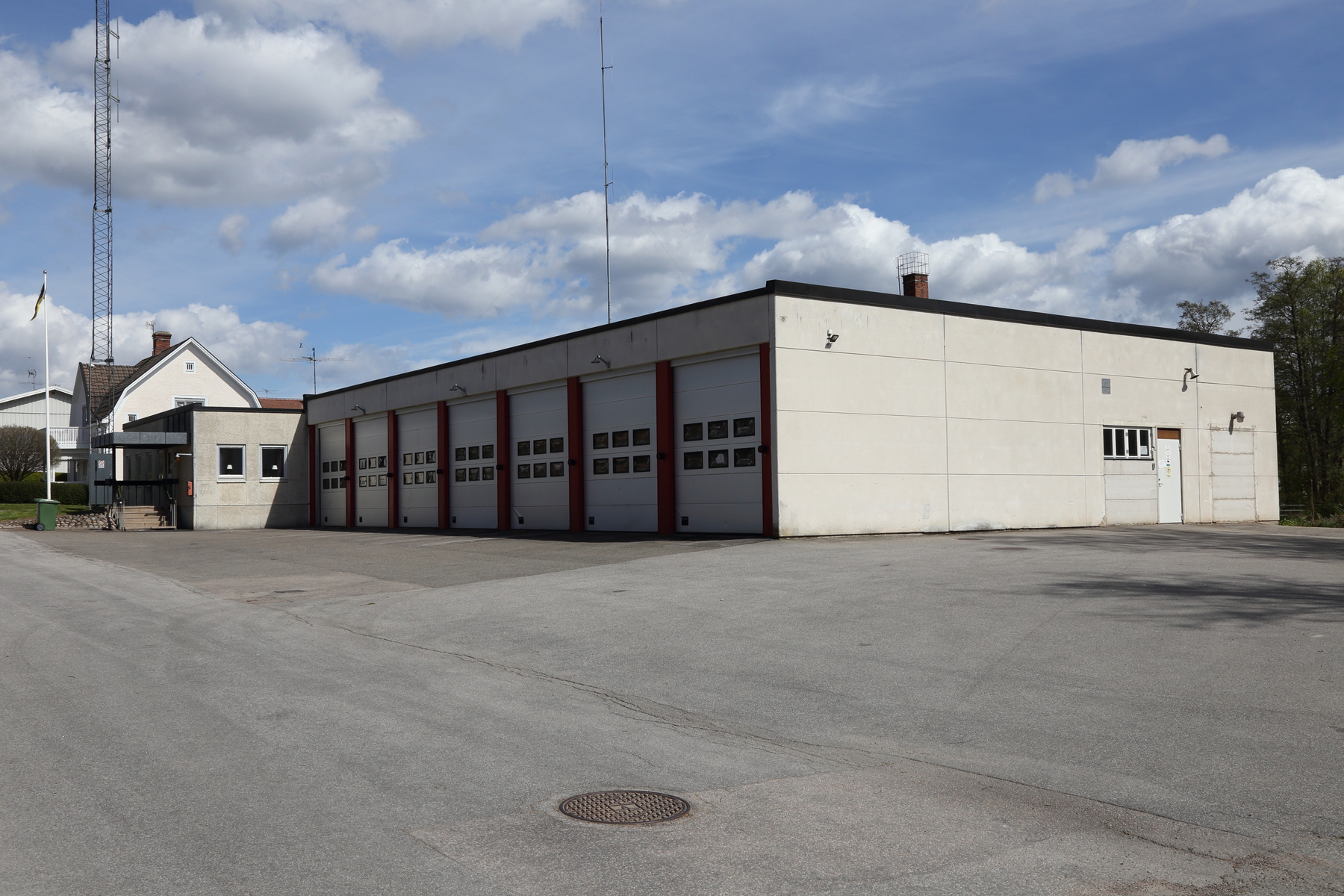 En modern byggnad med sex garageportar och en sidodörr är synlig under en delvis molnig himmel. Byggnaden har en ljus fasad och röda detaljer runt garageportarna. Framför byggnaden finns en asfalterad yta, i bakgrunden skymtar en radiomast och ett bo