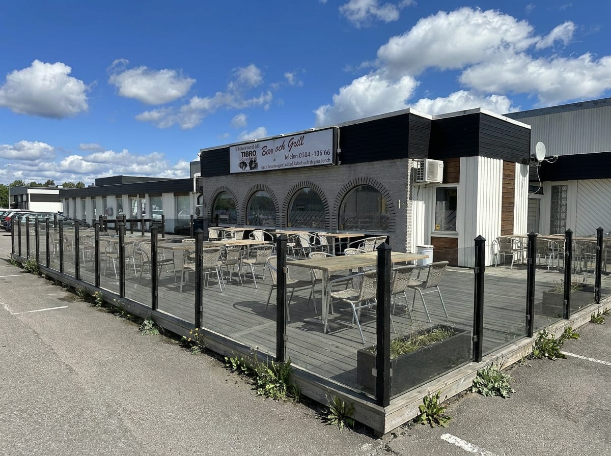 En utomhusbild som visar en restaurangbyggnad med en skylt märkt "TIBRO Bar och Grill". Utanför restaurangen finns en uteservering med trädäck, bord och stolar, omgiven av ett glasräcke. På marken runt uteserveringen är gräset grönt. Bakom restaurang