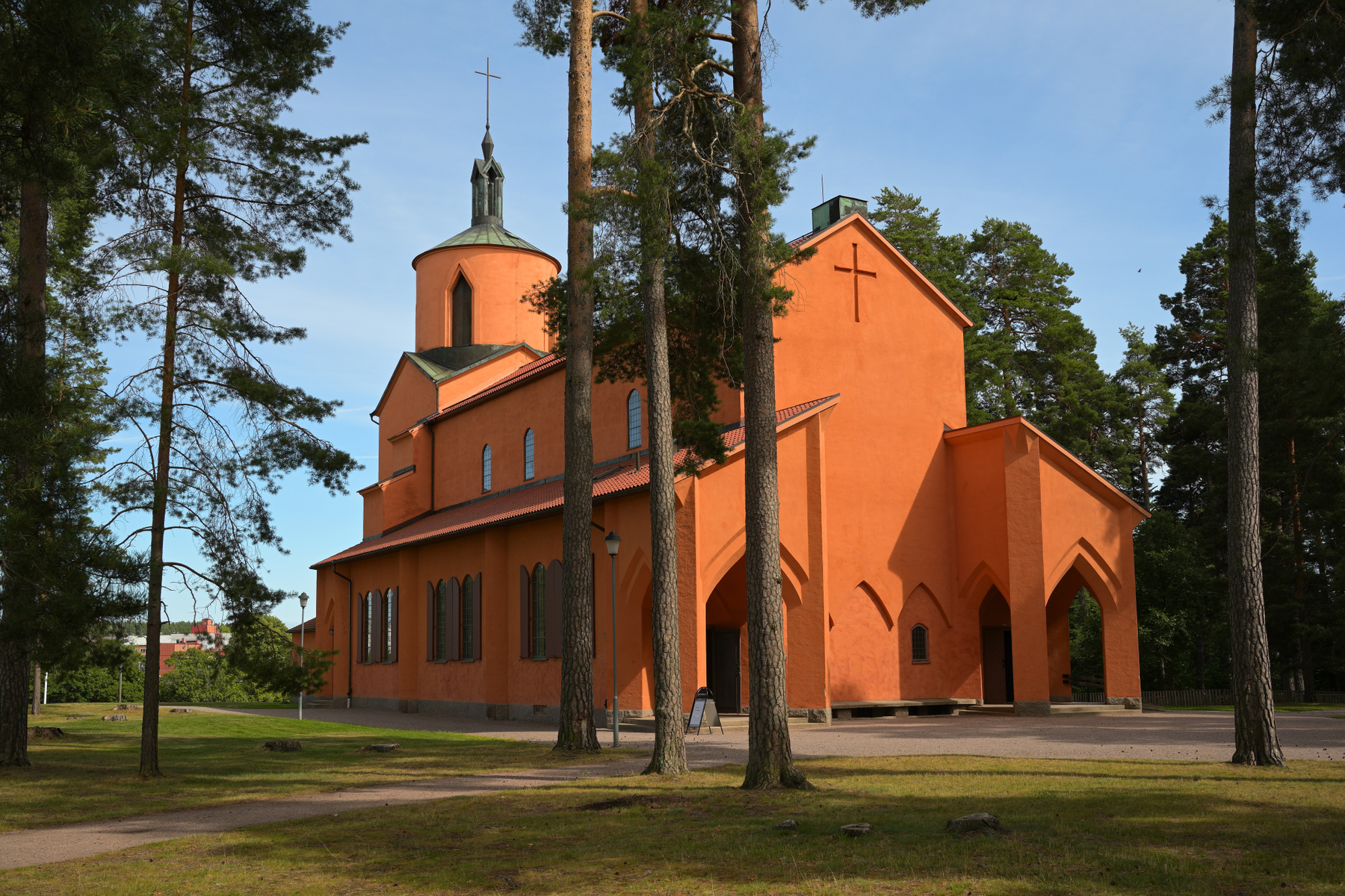 En stor, orange kyrkobyggnad med ett koppartak syns under en blå himmel, omgiven av gräsmattor och höga tallar. Kyrkan har en spetsig klocktorn med ett kors och flera valvformade fönster och dörröppningar. En grusgång leder förbi byggnaden.