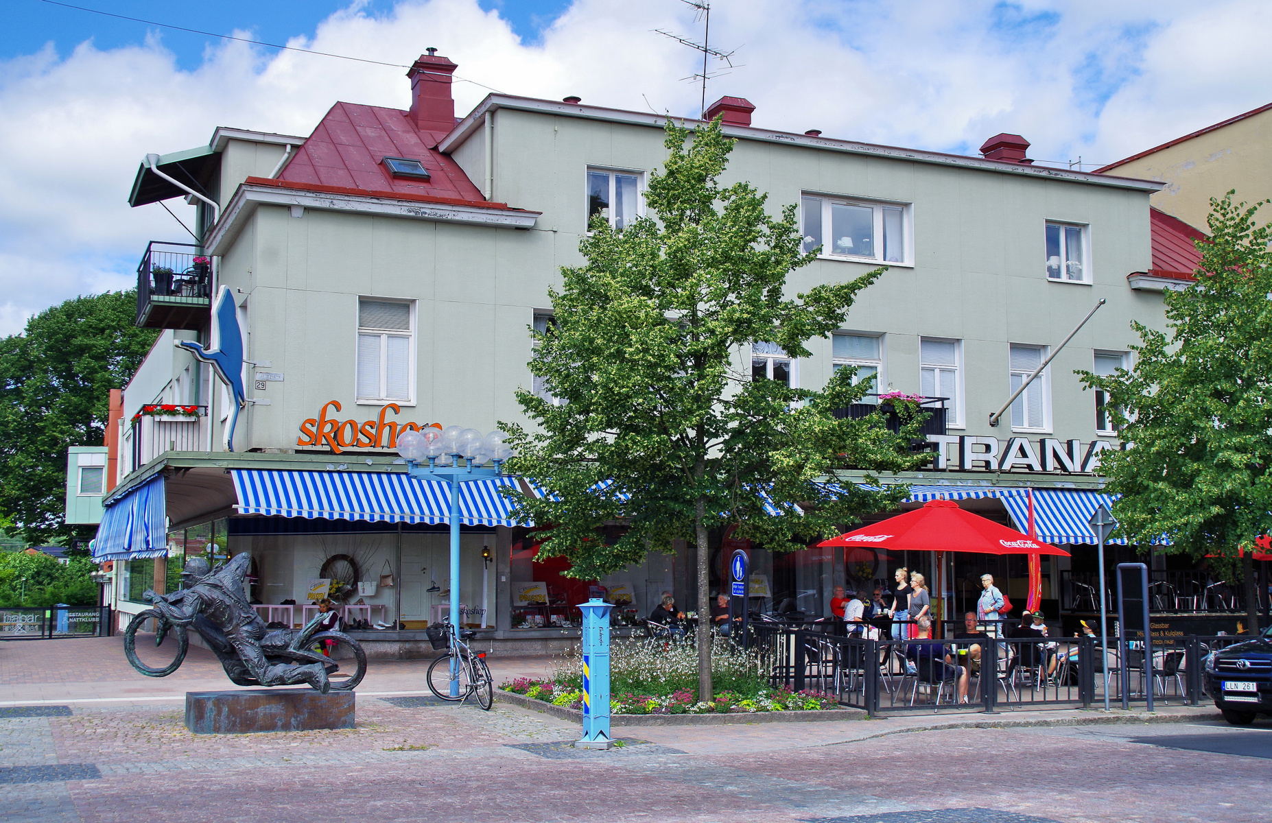 En bild visar en gata i en stad med en flervåningsbyggnad som inhyser affärer och restauranger på markplan. Framför byggnaden finns en terrass med bord och stolar under en röd parasoll, samt en staty föreställande en motorcyklist.
