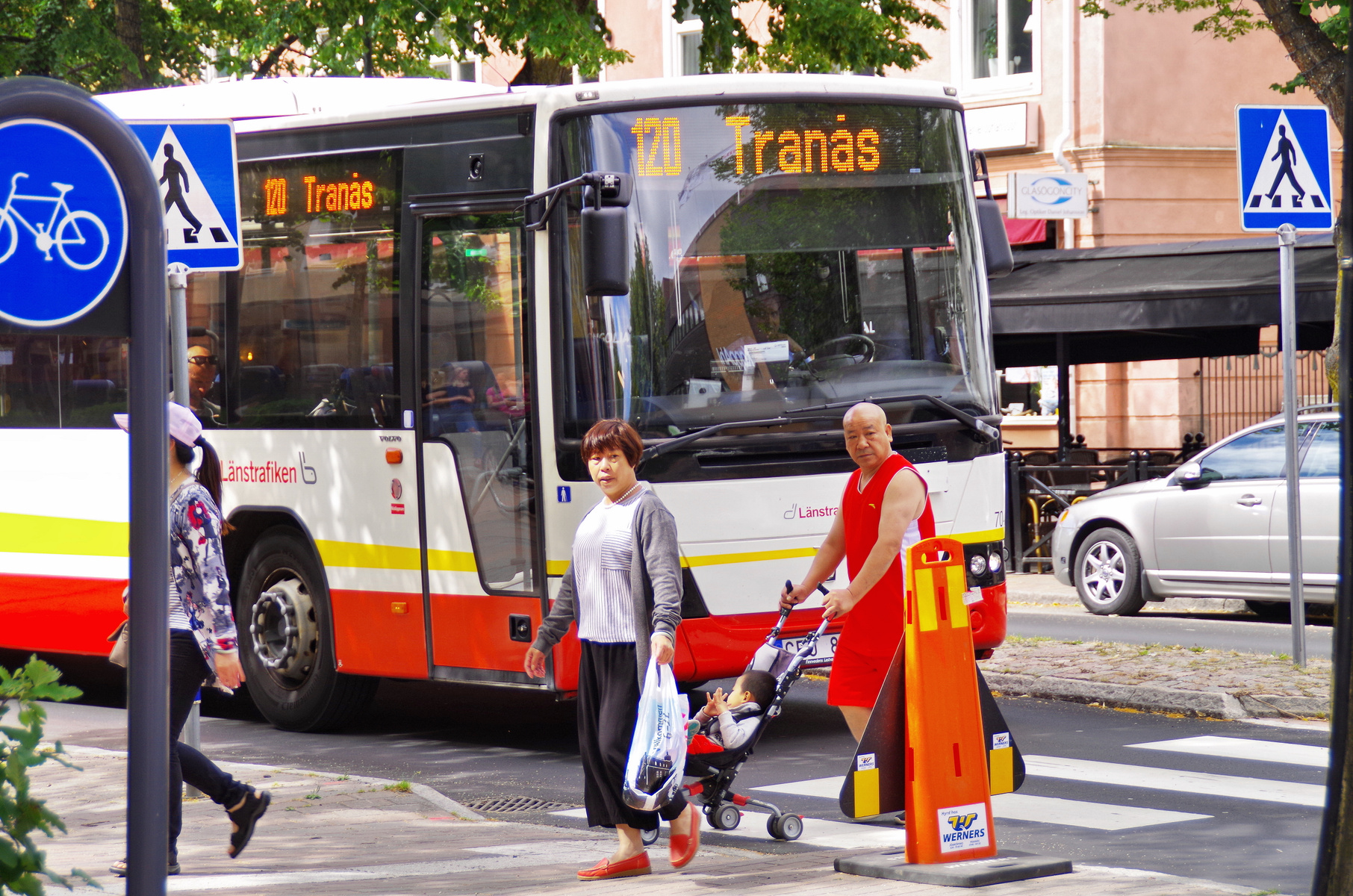 En gata med en buss som stannat vid ett övergångsställe där flera personer, inklusive en med en barnvagn, passerar. En kvinna håller en plastpåse. Två vägskyltar syns i förgrunden och en bil är parkerad vid sidan av gatan. Byggnader och träd utgör ba