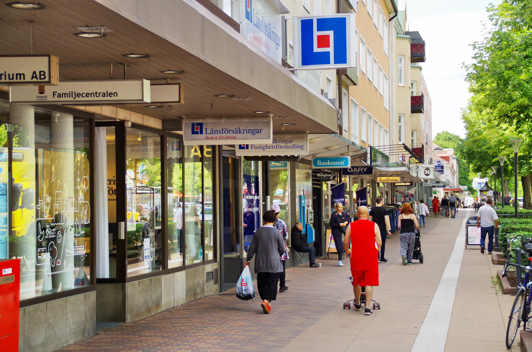 En gata med flera butiker och människor som går på trottoaren. Skyltar för "Länsförsäkringar", "Familjecentralen" och "Bankomat" syns. Det är en ljus dag med gröna träd längs gatan.