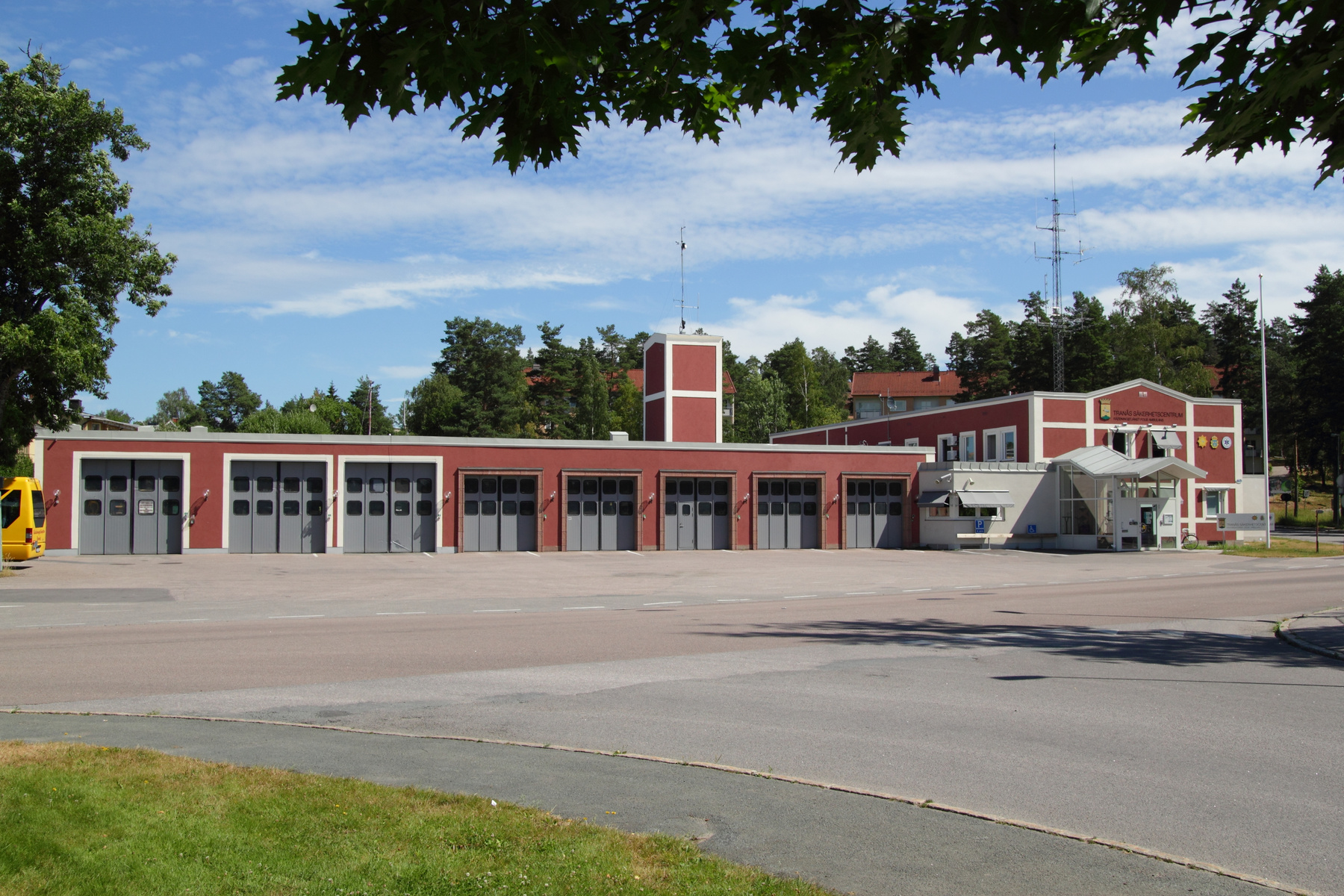 En röd byggnad med flera grå garagedörrar och vita detaljer under en klarblå himmel med vita moln. I bakgrunden syns träd och en mast. På byggnadens framsida står det "TRANÅS SÄKERHETSCENTRUM". En gul bilkropp är synlig vid en av garagedörrarna.