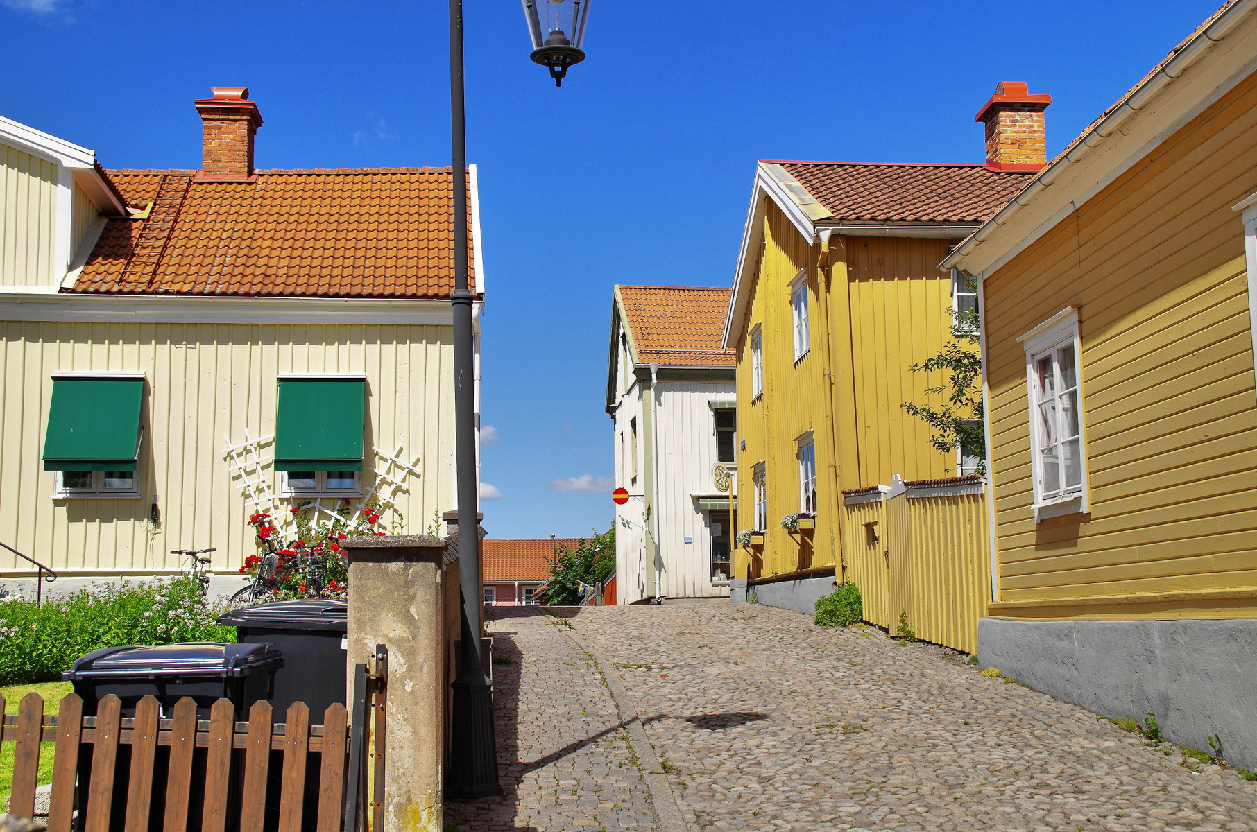 En smal, kullerstensbelagd gata omgärdad av trähus i ljusa färger som gult och gräddvitt under en klarblå himmel. På vänster sida syns ett hus med gröna fönstermarkiser och röda rosor. Ett staket, soptunnor och en lyktstolpe är synliga i förgrunden.