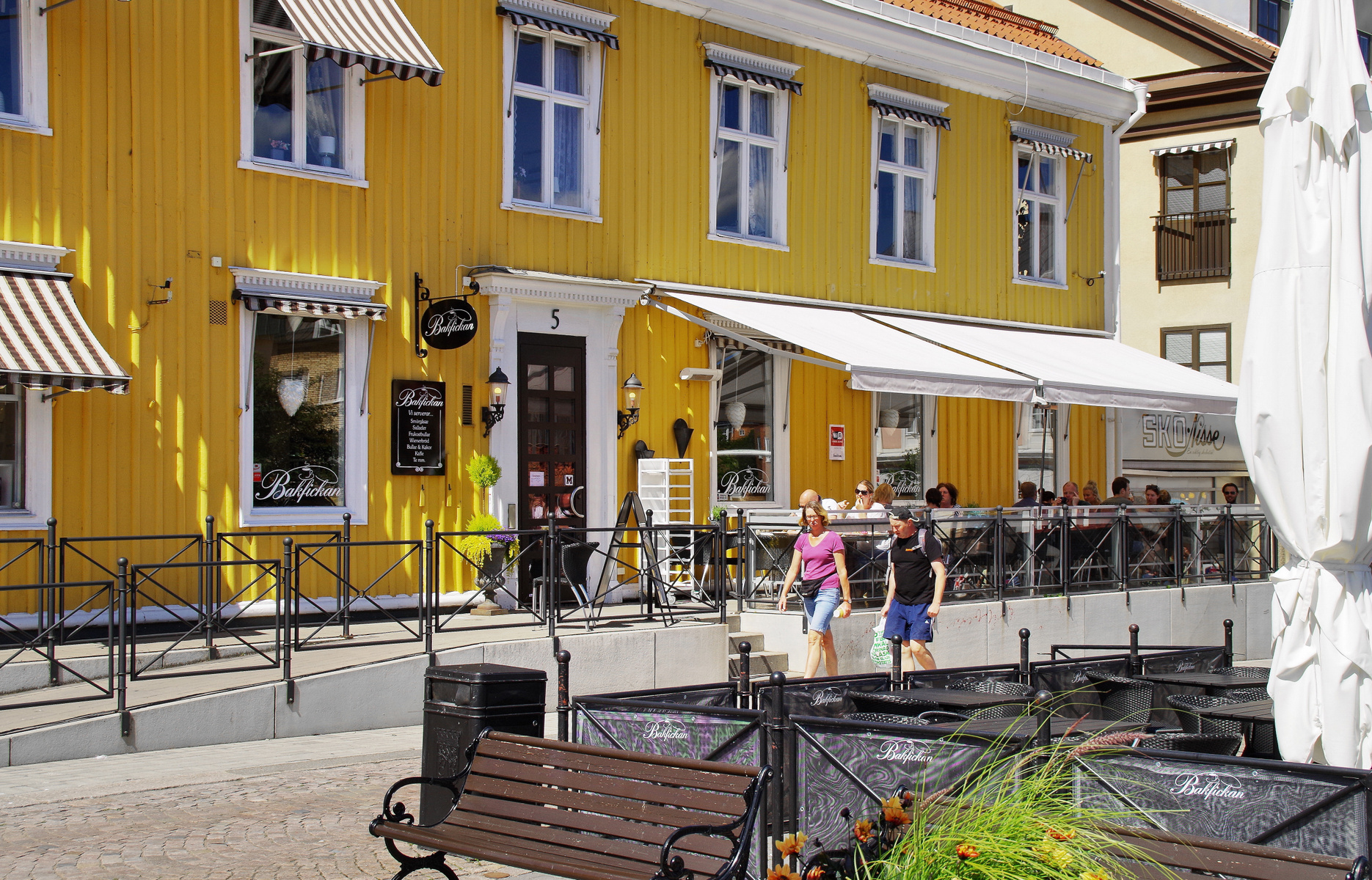 En gul trähusfasad med flera fönster och ränder på markiserna. Framför huset finns en terrass med sittplatser, delvis skuggad av markiser. Ett par personer går förbi terrassen, och ett par sitter på en bänk framför. På skyltar står "Bakfickan".