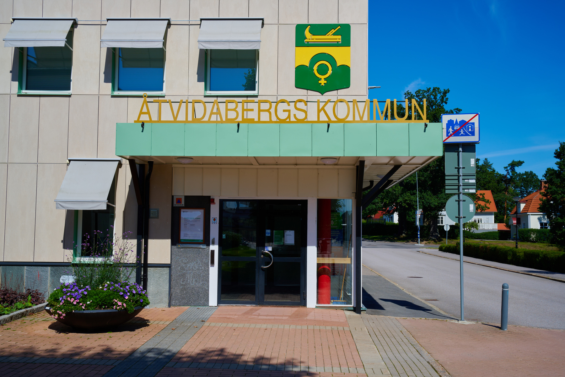 En fasad på en byggnad med skylten "Åtvidabergs Kommun" ovanför huvudentrén. En logotyp med grön bakgrund och gula symboler sitter vid sidan av ingången. Byggnaden har fönster med ljusa markiser och framför dörren står en blomlåda med lila och rosa b