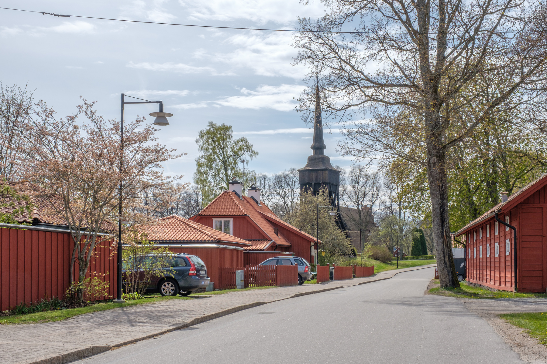 En väg kantad av röda trähus med tegeltak. Till vänster syns en parkerad personbil och en blommande buske. I bakgrunden reser sig en kyrkas spira. Träd, några med synliga knoppar och blad, ramar in bilden under en ljusblå, molnig himmel.
