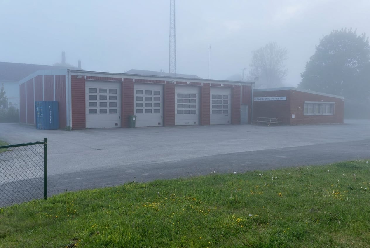 En röd byggnad med fyra vita garageportar och ett skyltfönster. Ett antenntorn syns i bakgrunden. Marken består av en asfalterad yta framför byggnaden och en gräsyta med maskrosor till vänster och i förgrunden. Allt är omslutet av dimma.