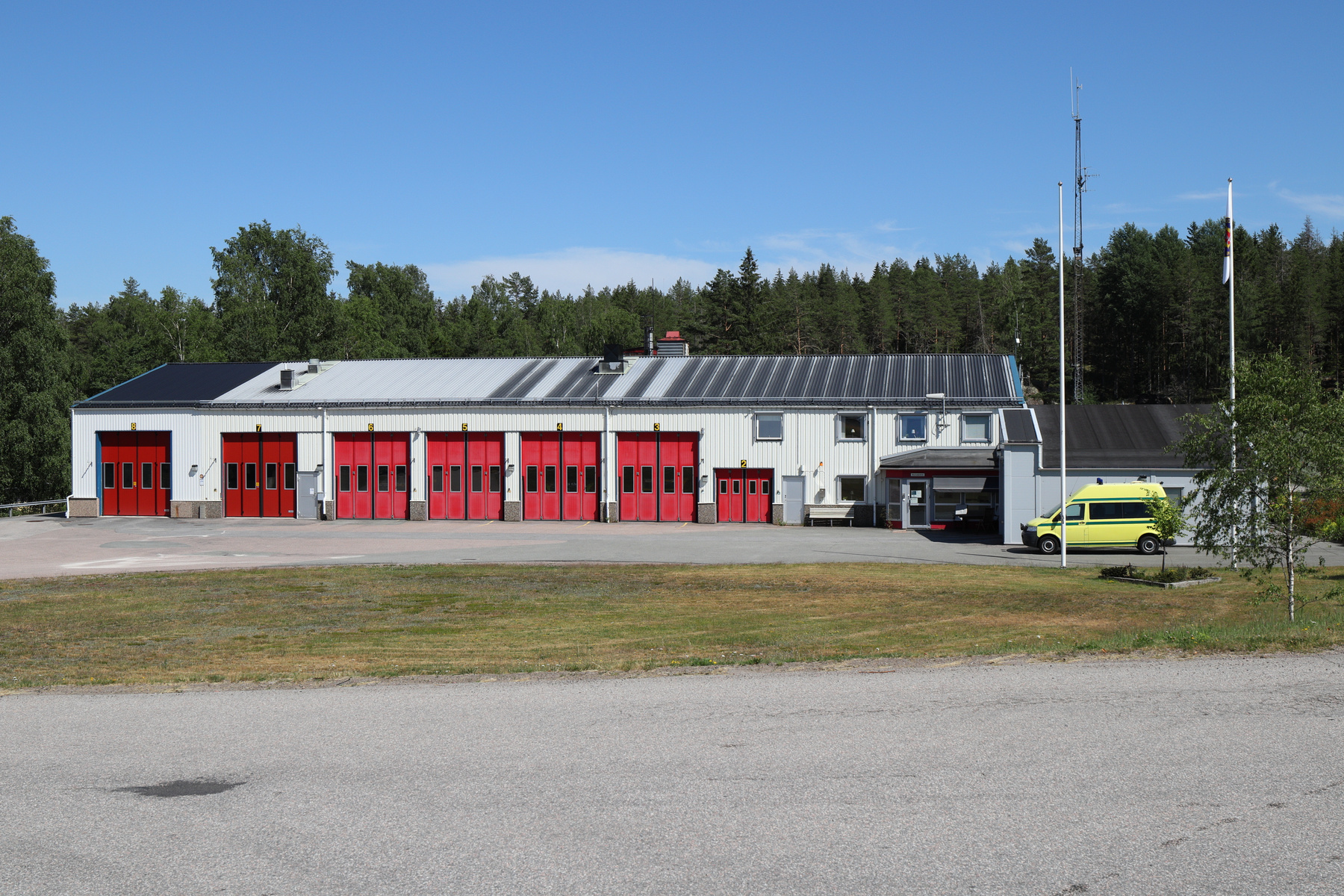 En enplansbyggnad med åtta röda garageportar och ett vitt plåttak som delvis är svart. Framför byggnaden finns en asfalterad yta, en gräsplätt och en väg. En gul bussfordon är parkerat vid byggnadens högra sida. I bakgrunden syns skog och en klarblå
