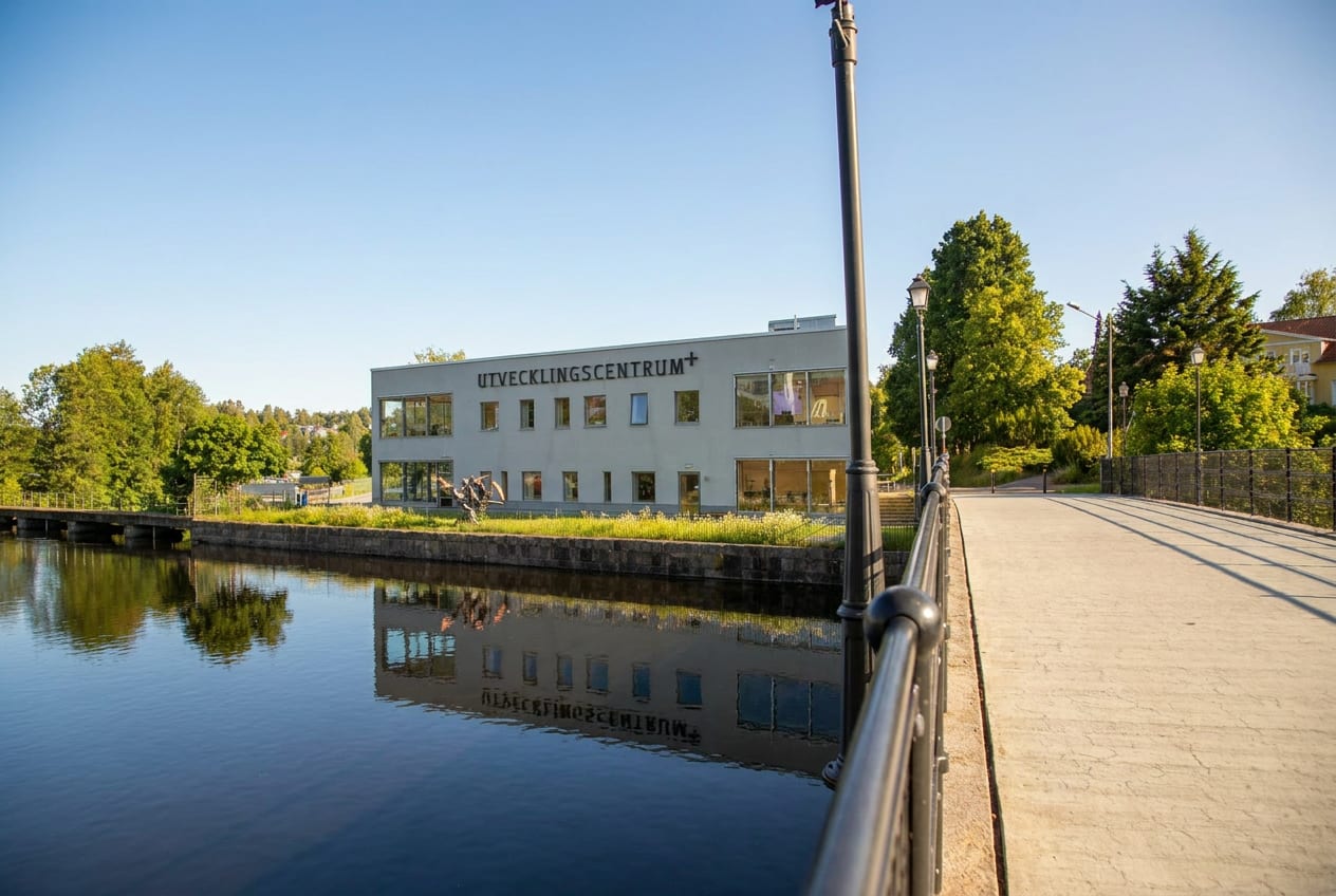 En modern byggnad med texten "UTVECKLINGSCENTRUM" på fasaden ligger vid en vattenyta som reflekterar byggnaden. Intill vattnet syns en bro med mörka räcken och gatumlyktor. Grönska ramar in miljö.