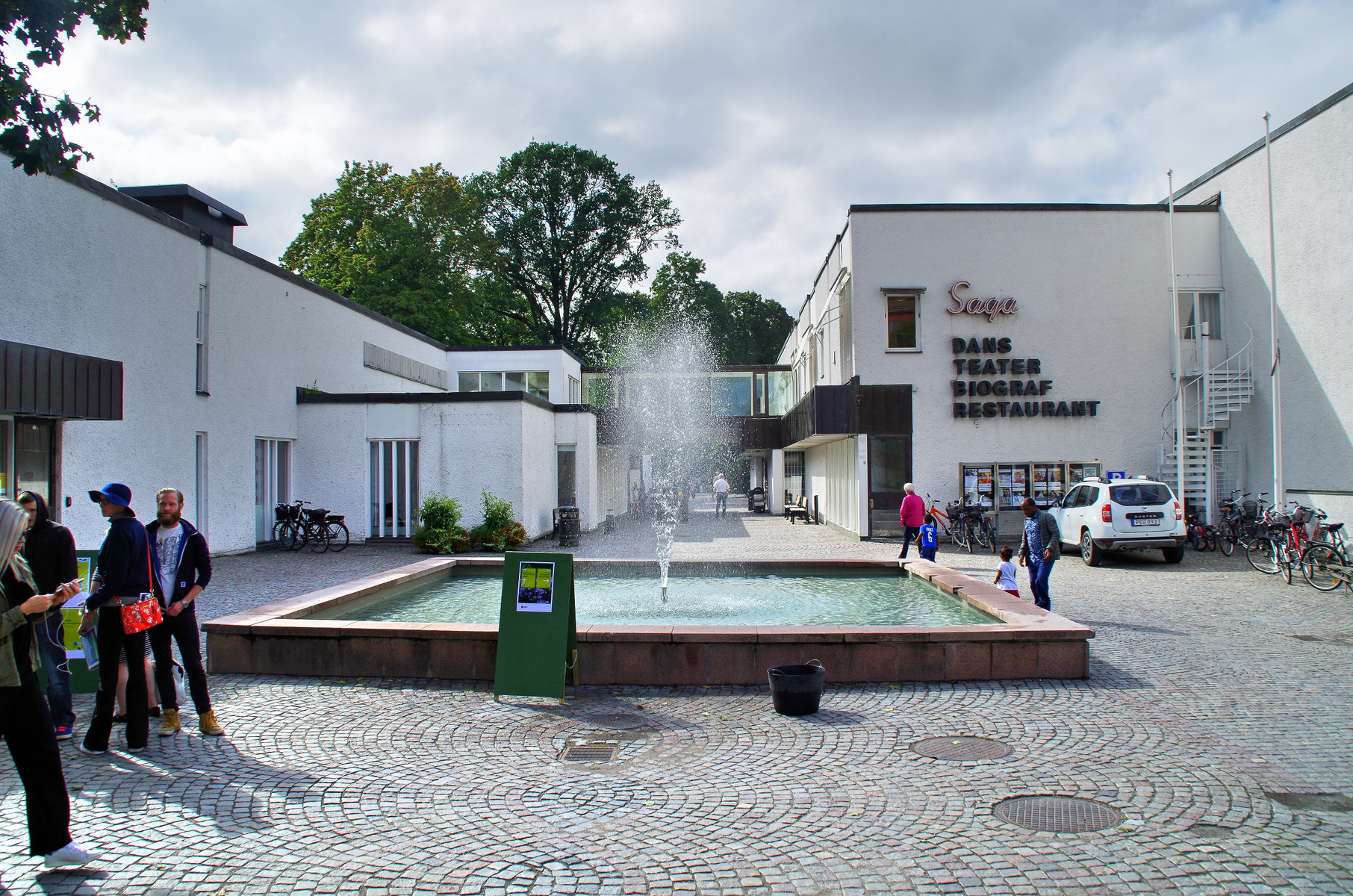 En utomhusbild som visar en modern vit byggnad med skylten "Saga Dans Teater Biograf Restaurant". Framför byggnaden finns ett stenbelagt torg med en rektangulär fontän där vatten sprutar upp. Flera personer, cyklar och en vit bil kan ses på torget.