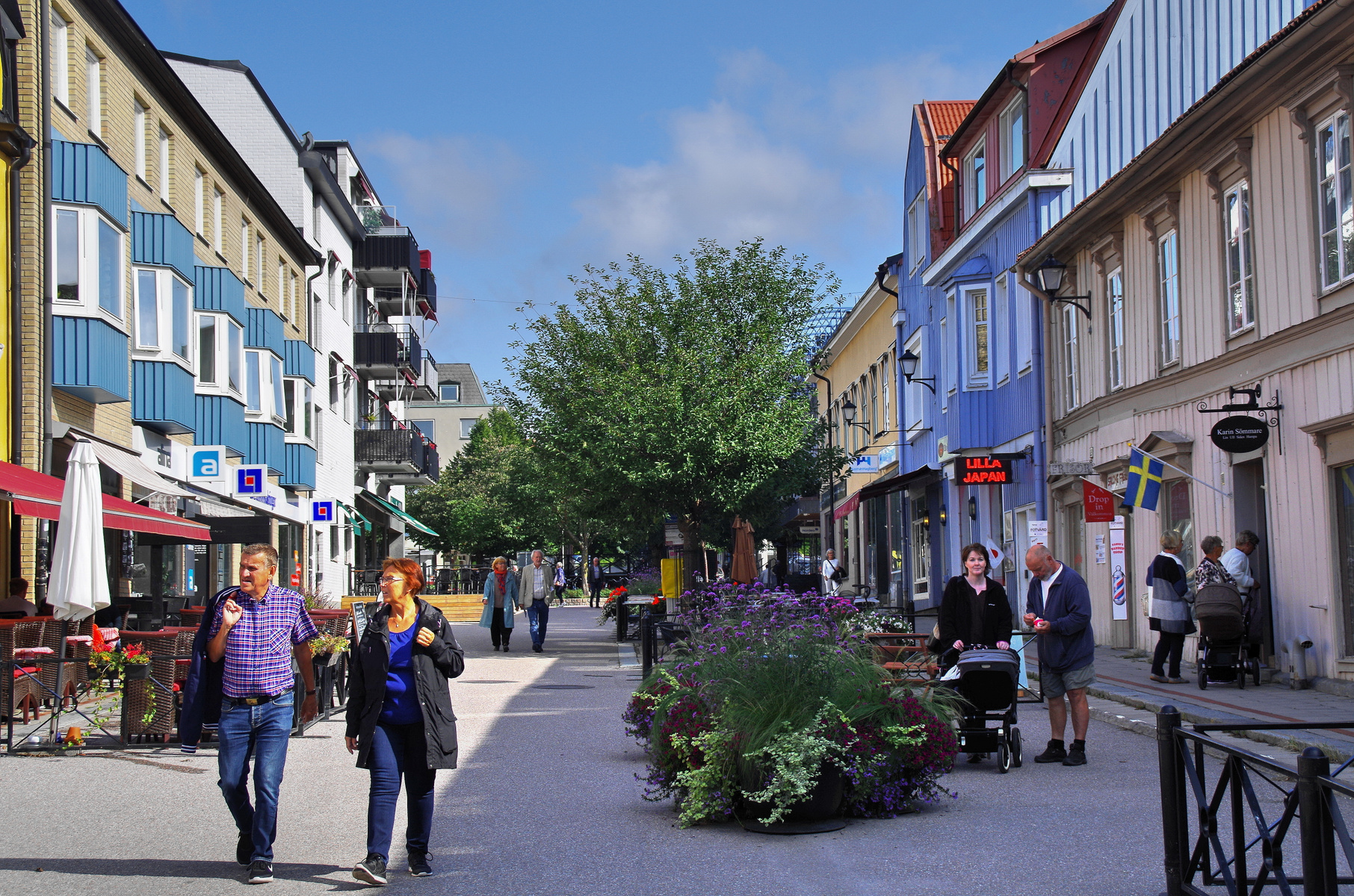 En gata kantad av flervåningshus med butiker och restauranger på bottenvåningen. Flera människor syns gående på gatan som är dekorerad med planteringar. Blå himmel med vita moln är synlig ovanför husen.