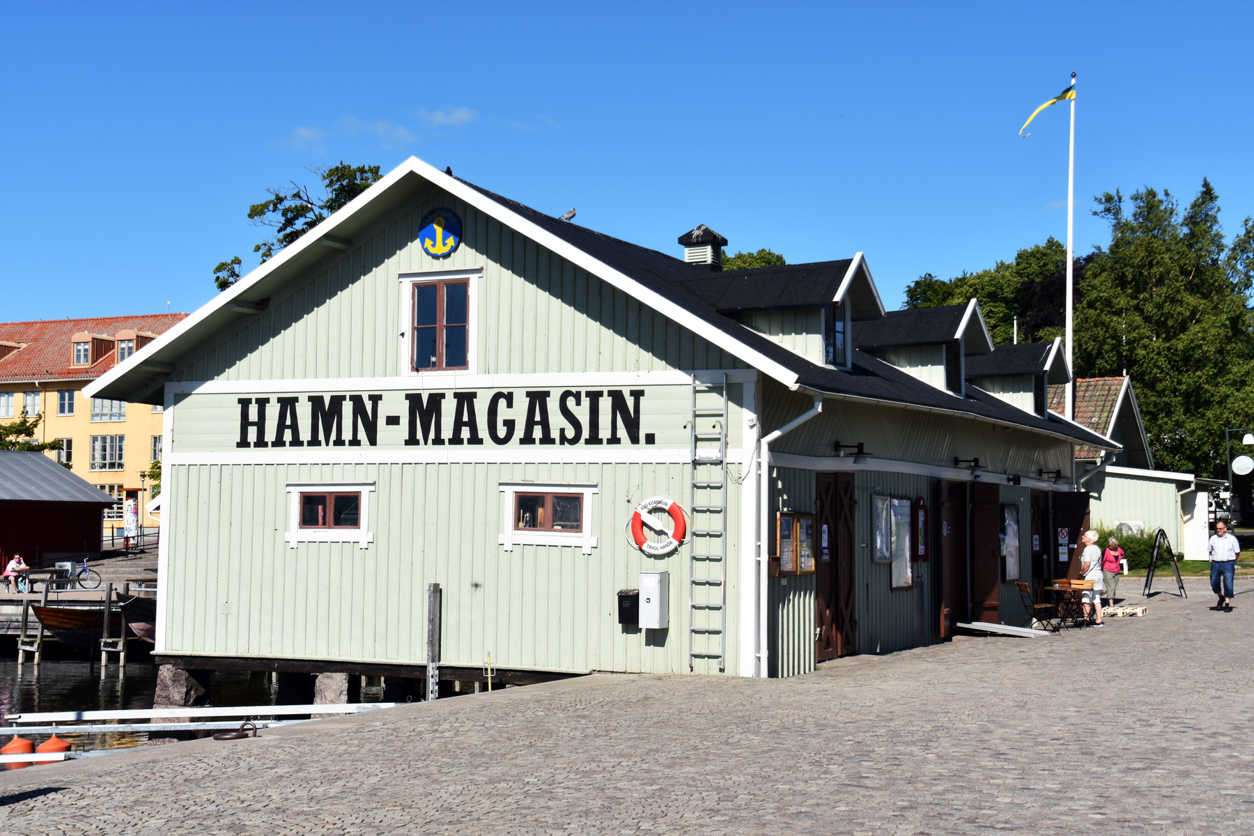 En ljusgrön träbyggnad med texten "HAMN-MAGASIN." på fasaden står vid en brygga. Till höger om byggnaden finns en gatstensbelagd yta med människor och en flaggstång med en gul vimpel. I bakgrunden syns andra byggnader, träd och blå himmel.