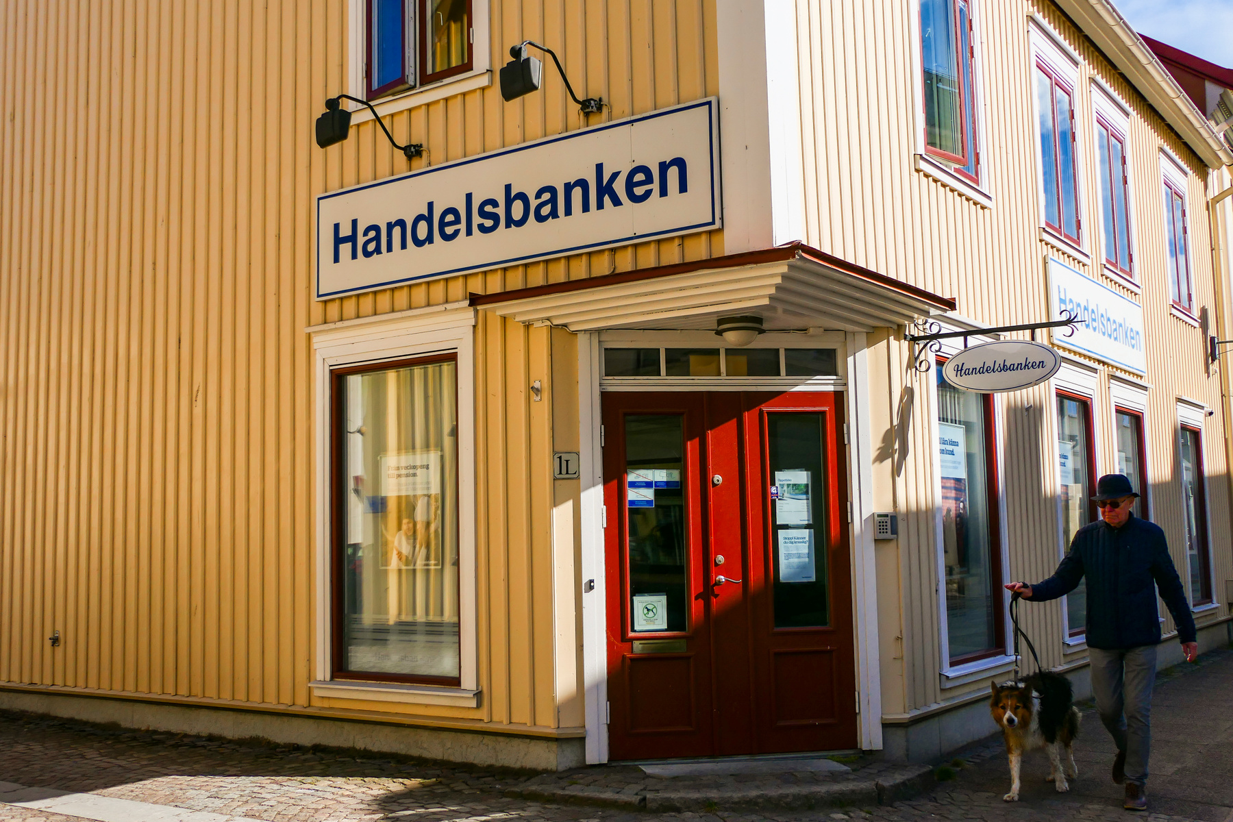 En gul byggnad med röd dörr och skyltar som visar "Handelsbanken". En person med hatt går förbi med en hund i koppel på en kullerstensbelagd trottoar. Solen lyser på byggnaden och skapar skuggor.
