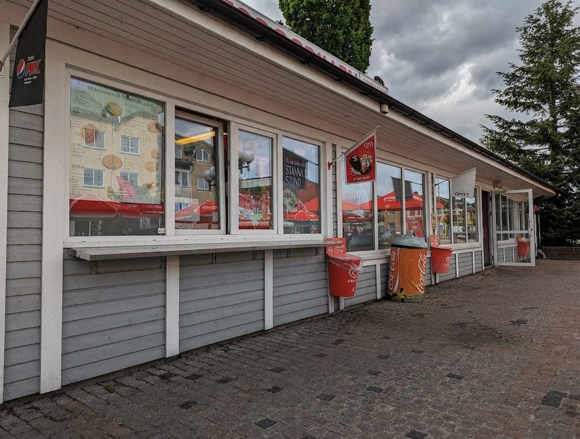 En enplansbyggnad med grå träfasad och vita knutar, vars stora fönster reflekterar röda parasoller. Framför byggnaden står röda soptunnor och en orange burkformad behållare. Det är en molnig dag och marken är täckt av grå marksten.