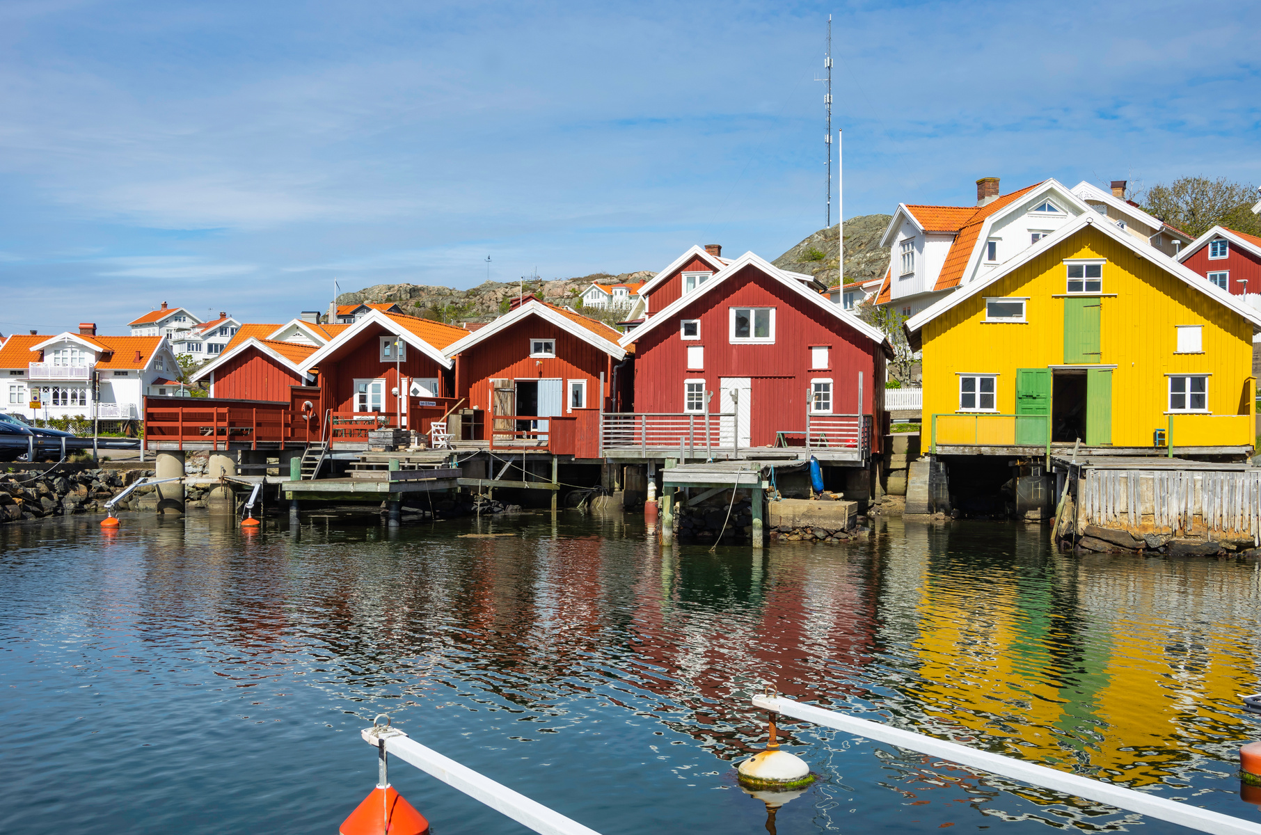 Bilden visar en rad med sjöbodar i olika färger, främst röda och en gul, som står på pålar över vatten med sina reflektioner synliga. I bakgrunden syns bostadshus, en kulle och en mast. Två båtbojar flyter i vattnet i förgrunden.