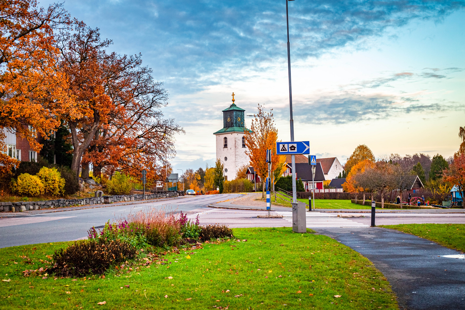 En gatuscen från ett bostadsområde under hösten, med träd i klara höstfärger och en kyrka med en grön kupol och klocka i bakgrunden. Ett övergångsställe syns i förgrunden och vägskyltar står vid sidan av vägen.