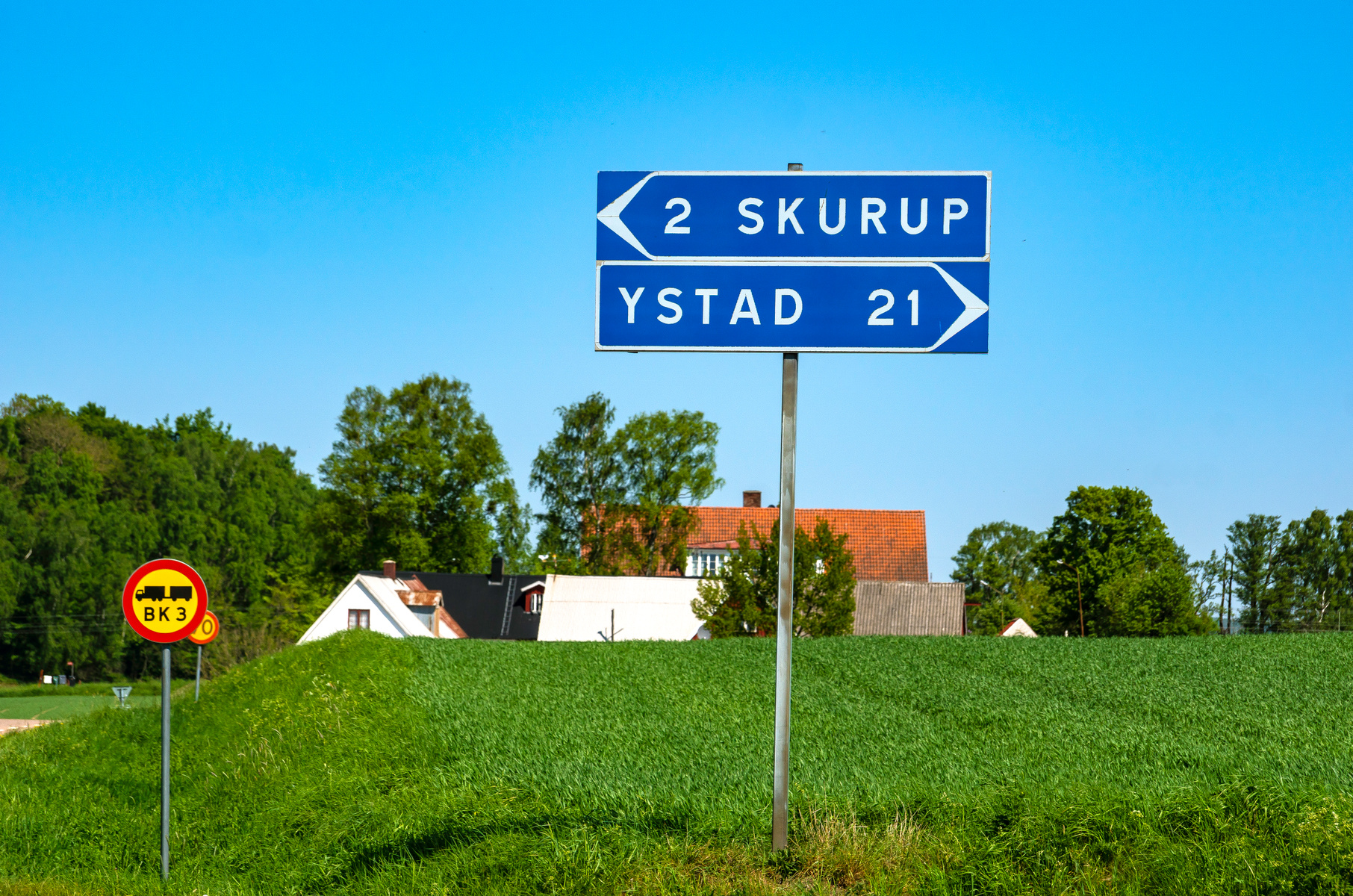 En blå vägskylt med vit text som visar riktning mot Skurup (2 km) och Ystad (21 km). En varningsskylt med en lastbilssymbol och texten "BK 3" syns också. Bakom skyltarna ligger ett grönt fält, träd och hustak under en ljusblå himmel.