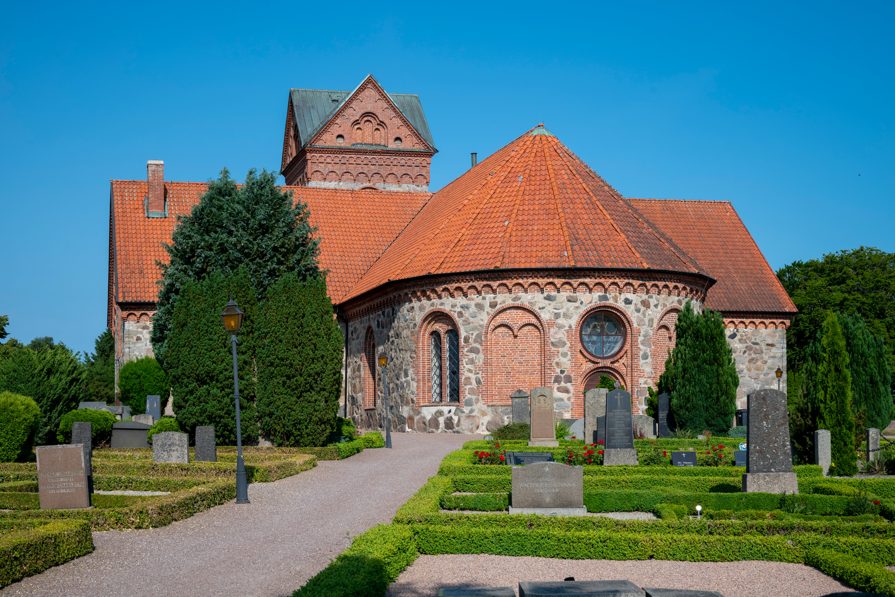 En kyrka med tegel- och stenfasad samt rött tegeltak under en klarblå himmel. På kyrkogården syns grusgångar, klippta häckar, träd, buskar, gravstenar och en lyktstolpe.