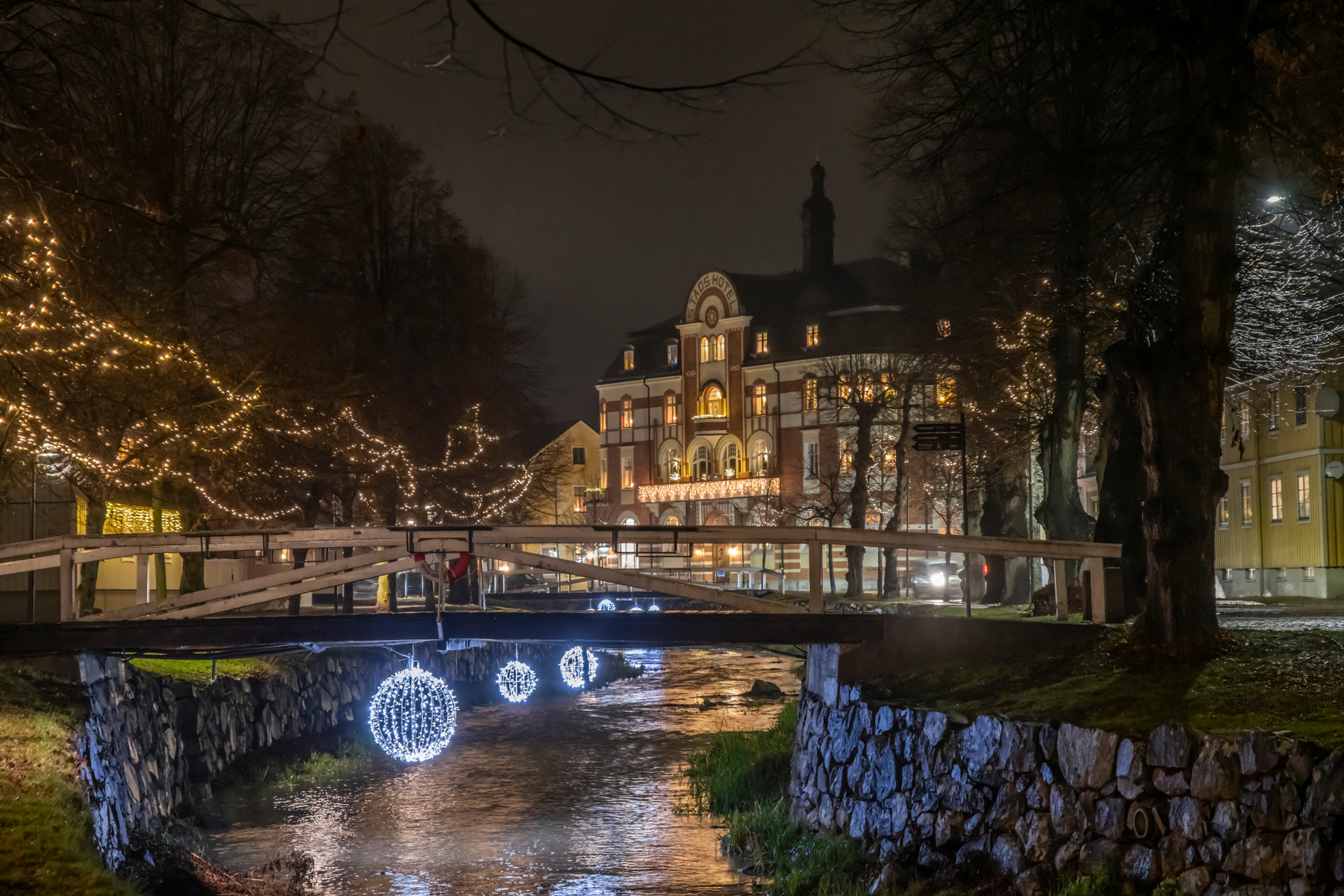 En nattbild som visar en upplyst stadsmiljö med en kanal eller å i förgrunden. En bro går över vattnet, och ett större byggnad med fasadutsmyckning syns i bakgrunden. Träd längs vattnet och vid byggnaden är dekorerade med ljusslingor, och runda ljusd