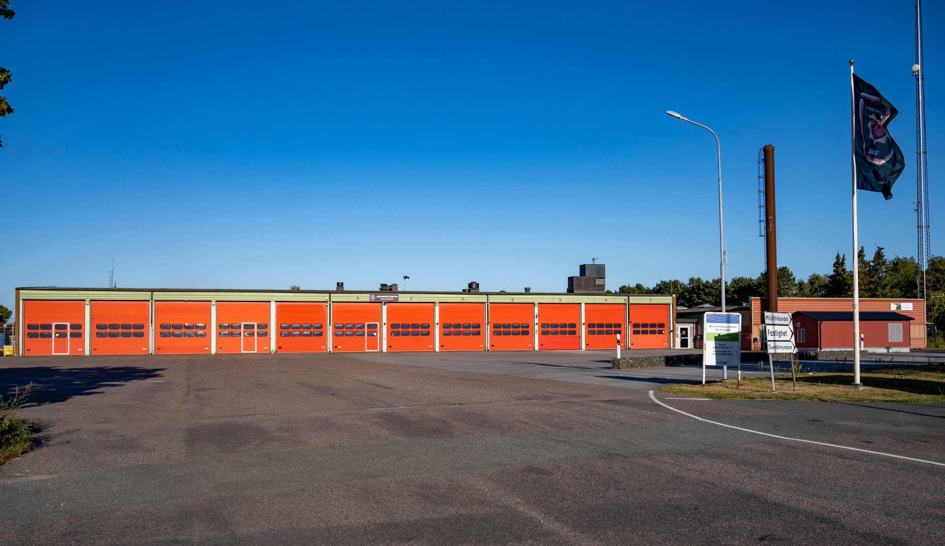 En byggnad med orangea portar under en klarblå himmel. Framför byggnaden finns asfalterad mark och till höger en flaggstång med en mörk flagga, en ljusstolpe och ett brunt torn. Några skyltar står vid infarten och träd syns i bakgrunden.