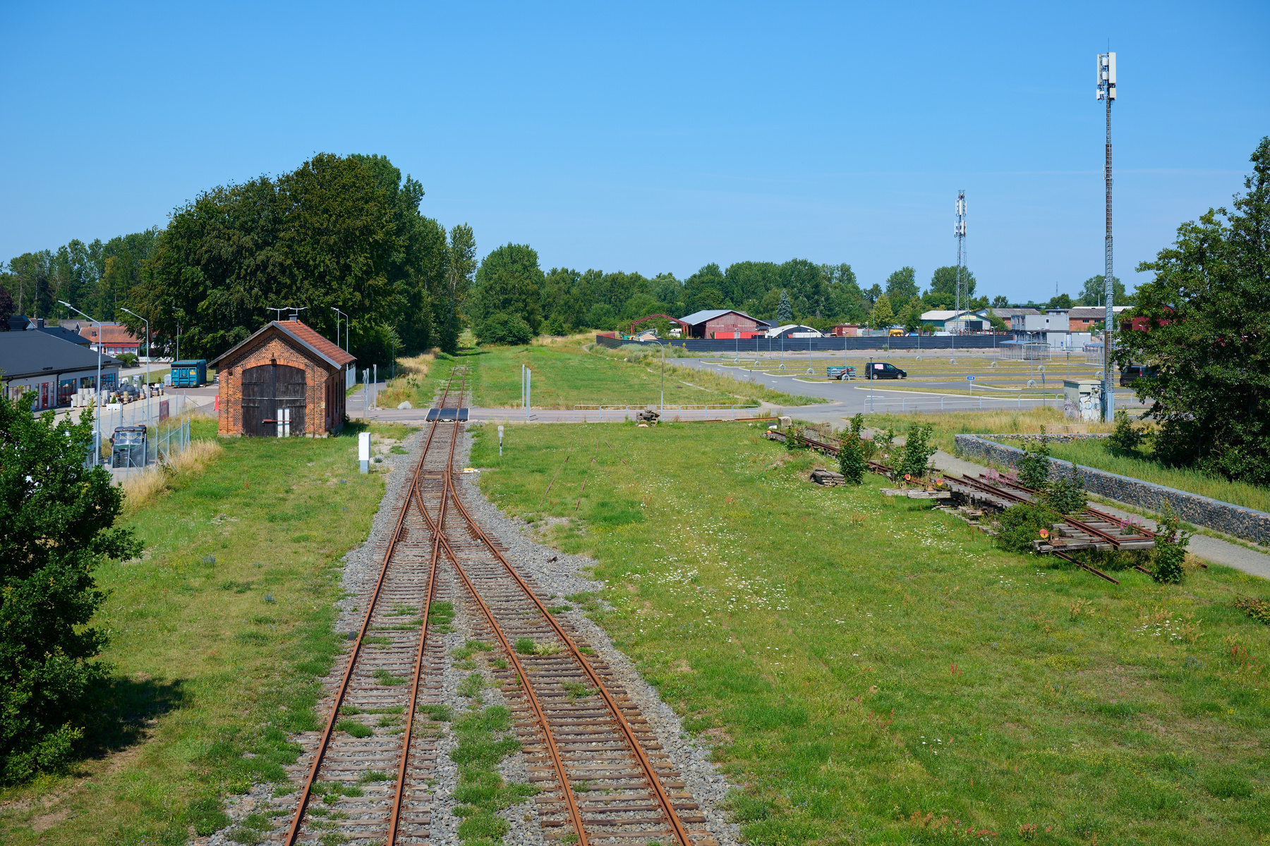 En översiktsbild av ett område med flera järnvägsspår som strålar ut från förgrunden. I mitten ses en tegelbyggnad med stora dubbla trädörrar, och i bakgrunden syns andra byggnader, träd, ett parkeringsområde med bilar samt en hög mast.