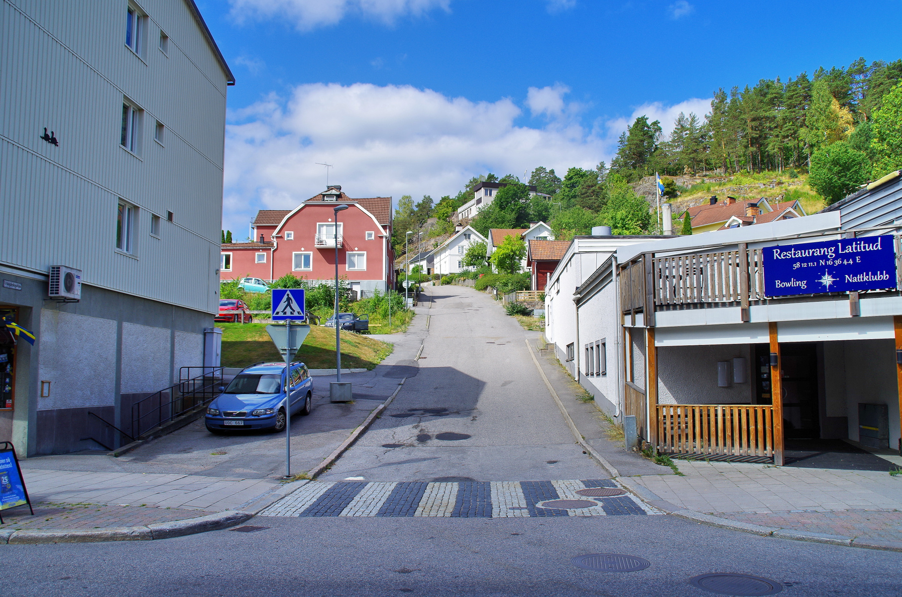 En gata framför en byggnad med en blå bil parkerad vid trottoaren. Till höger syns en byggnad med en skylt som anger "Restaurang Latitud". Längre bort syns en backe med hus och träd. En fotgängarskylt och ett övergångsställe syns i förgrunden.
