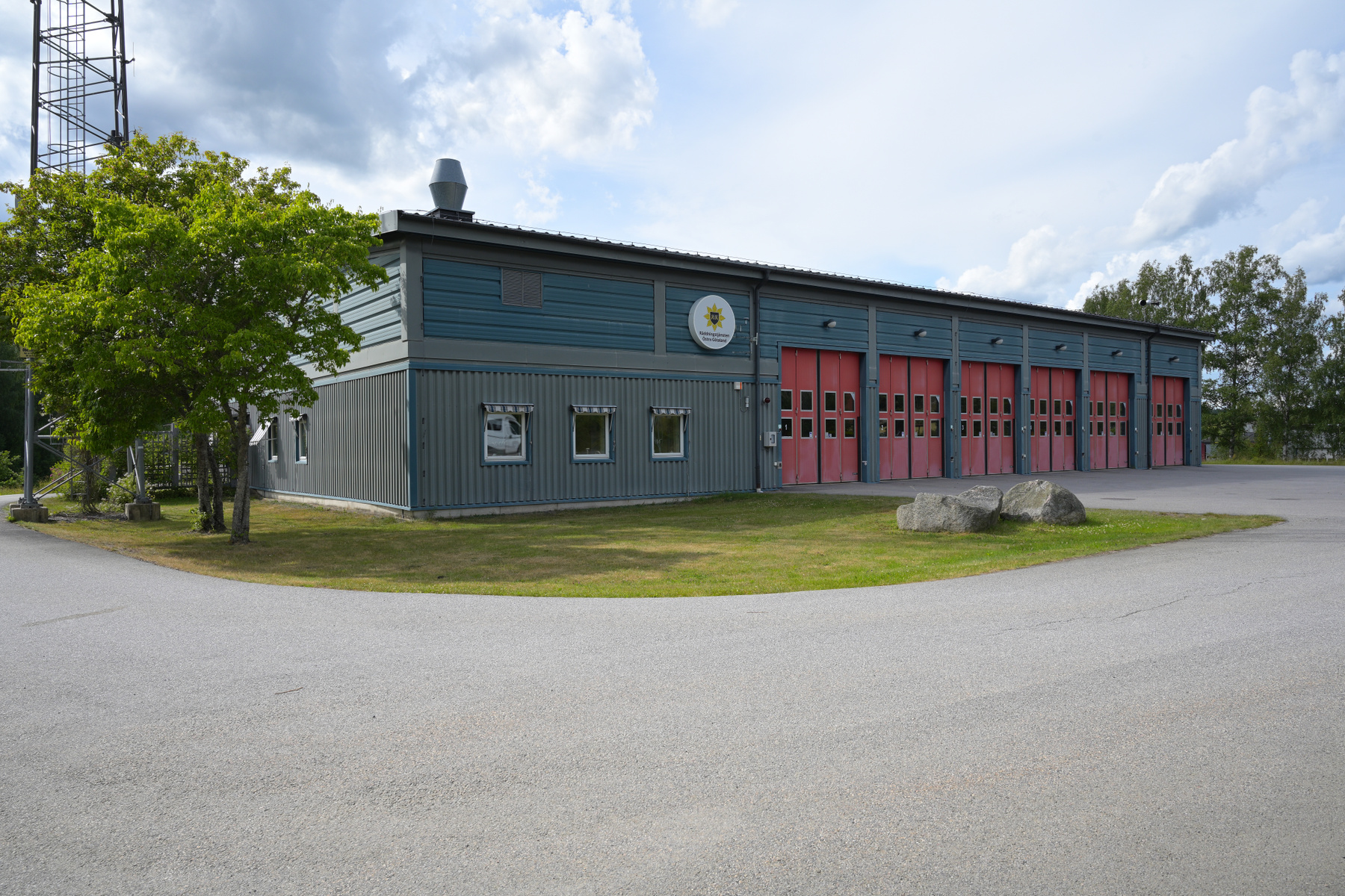 En modern byggnad med grå väggar och ett grått tak. På framsidan finns flera röda portar samt fönster. Byggnaden är omgiven av gräsmatta, en asfalterad yta och träd. En skylt med texten 'Räddningstjänsten Östra Götaland' är synlig på fasaden.