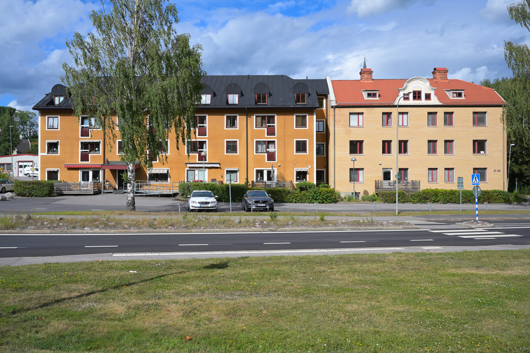 En bild tagen utomhus under dagtid som visar en gräsyta, en asfaltsväg med vita markeringar och i bakgrunden två flerfamiljshus i olika färger, det ena gult och det andra tegelfärgat. Flera bilar är parkerade framför husen.