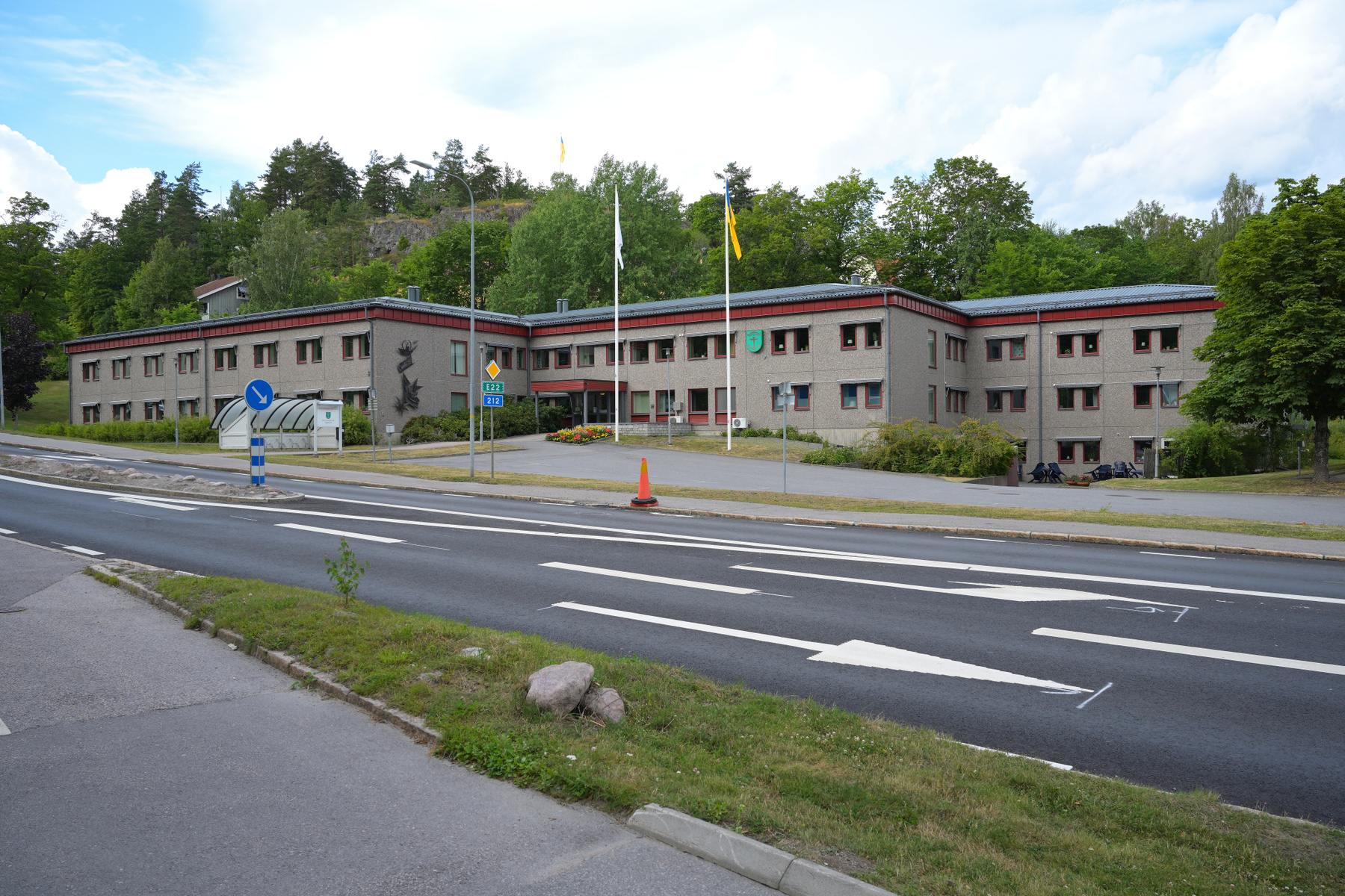 En tvåvåningsbyggnad med grå fasad och röda detaljer står intill en väg med vita pilar och markeringar. Grönskande träd och buskar omger byggnaden och en kulle syns i bakgrunden. Två flaggstänger med flaggor är placerade framför byggnaden.