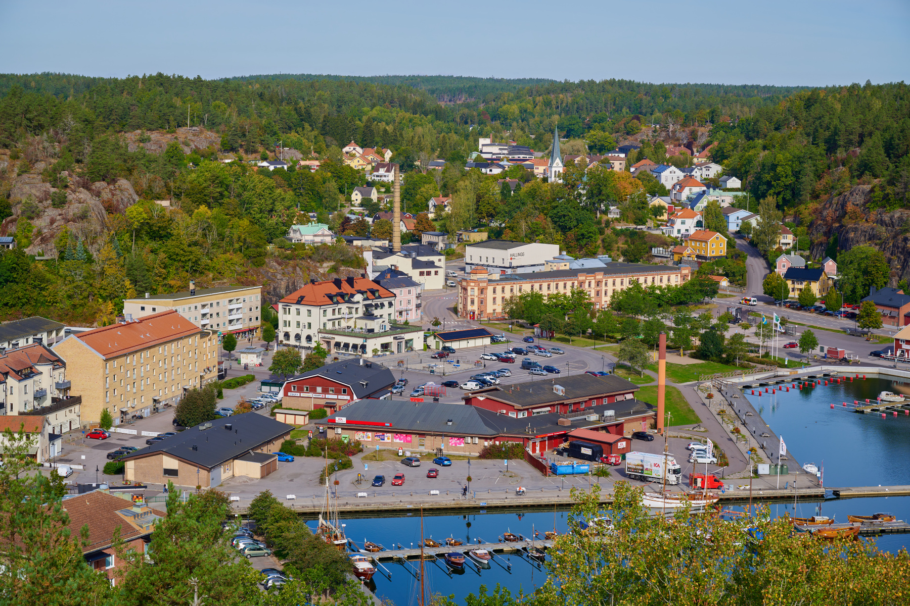 En översiktsbild av en stad som gränsar till vatten. Staden har en blandning av bostadshus, kommersiella byggnader och en kyrka, omgiven av grönskande kullar och träd. En hamn med båtar ligger vid vattnet.