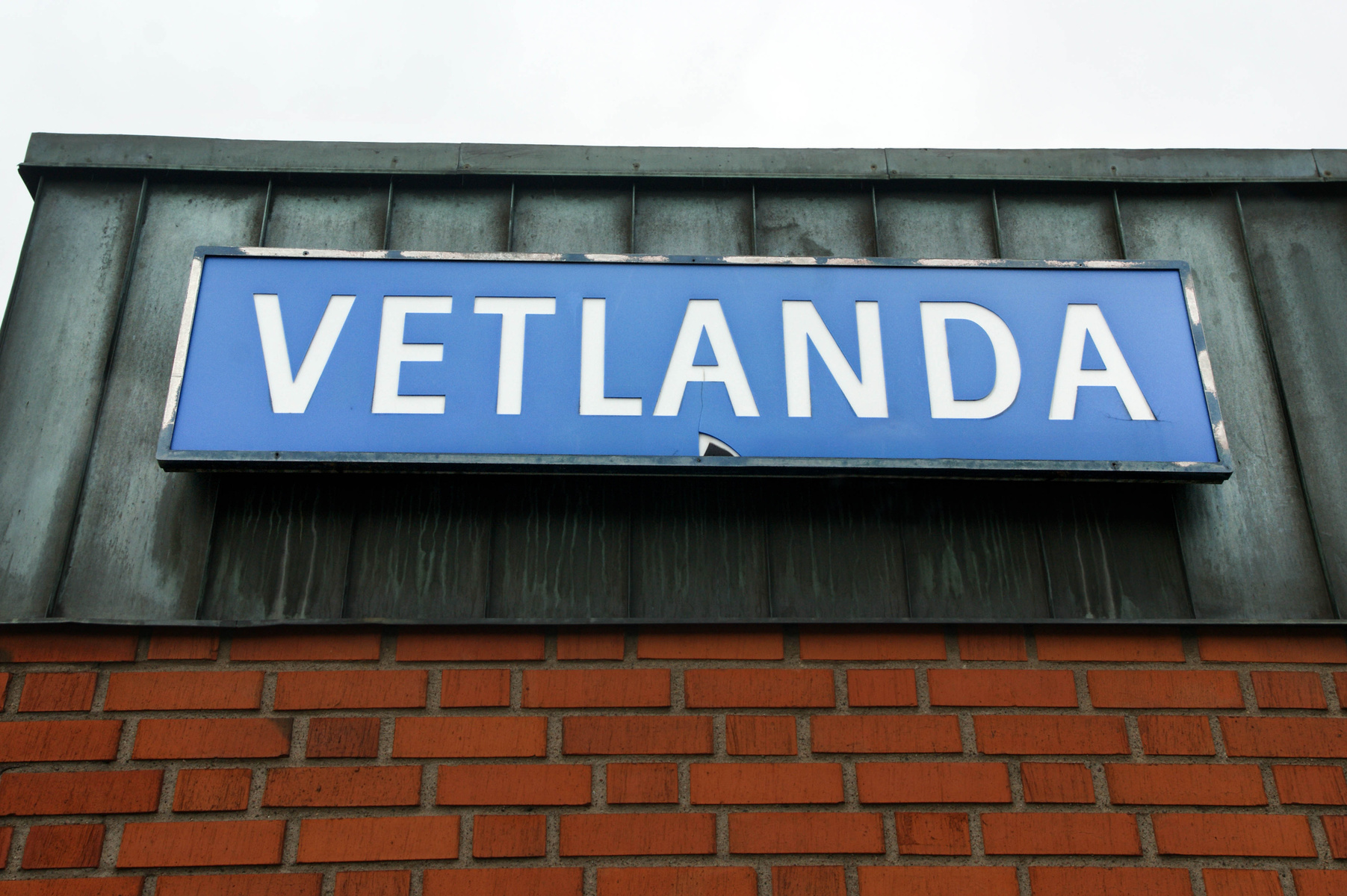 En blå skylt med vita bokstäver där texten "VETLANDA" står är monterad under en mörk takfot. Skylten hänger ovanför en tegelvägg. Himmel syns i bakgrunden.