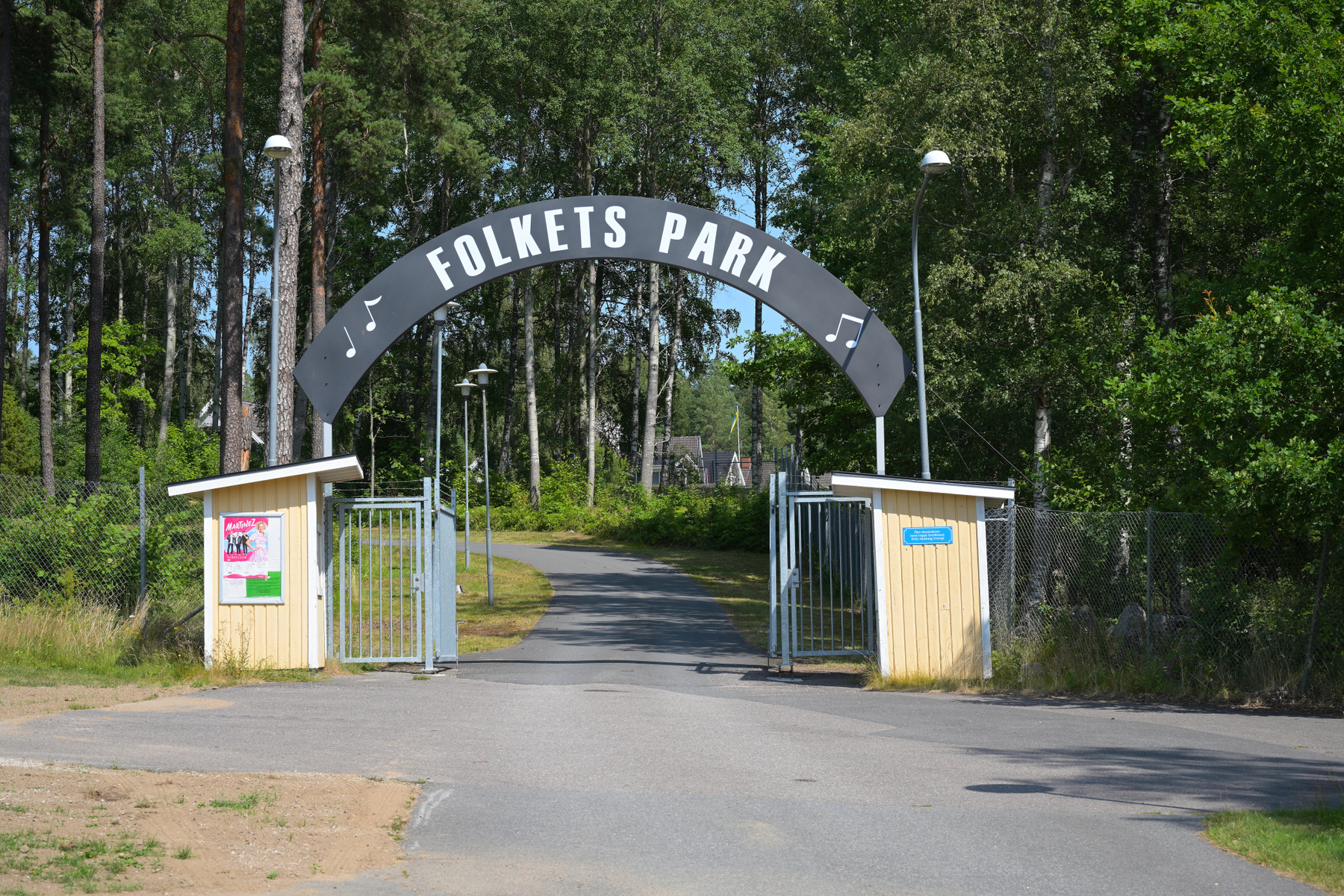 En entré med en skylt som det står "FOLKETS PARK" på. Skylten är bågformad och svart med vita bokstäver och två vita notsymboler. På båda sidor om grinden finns små gula byggnader med fönster och tak. Innanför grinden leder en asfalterad väg upp mot