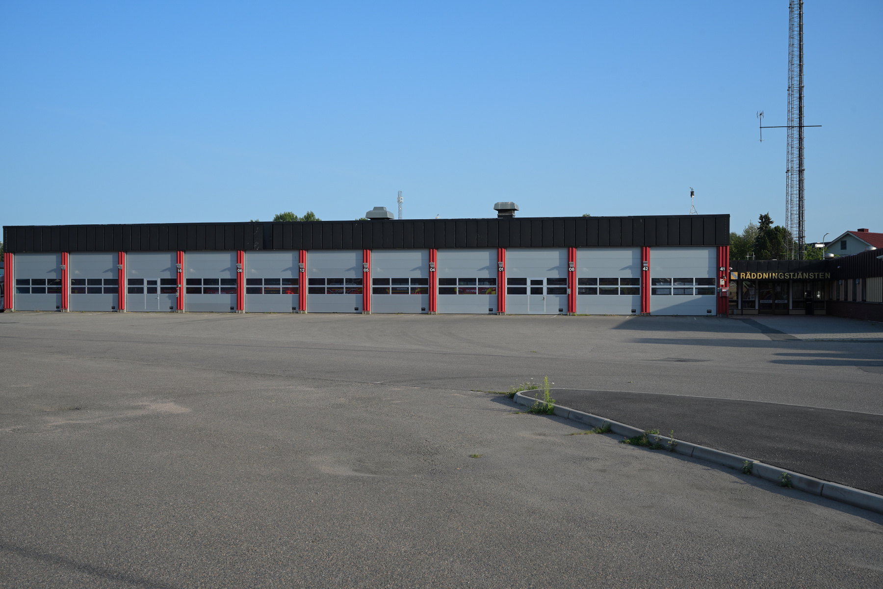 En bild som visar framsidan av en byggnad med flera garageportar i två våningar. Utanför byggnaden finns ett asfalterat område och i bakgrunden syns skog och en mast. På höger sida av byggnaden finns en skylt med texten RÄDDNINGSTJÄNSTEN.