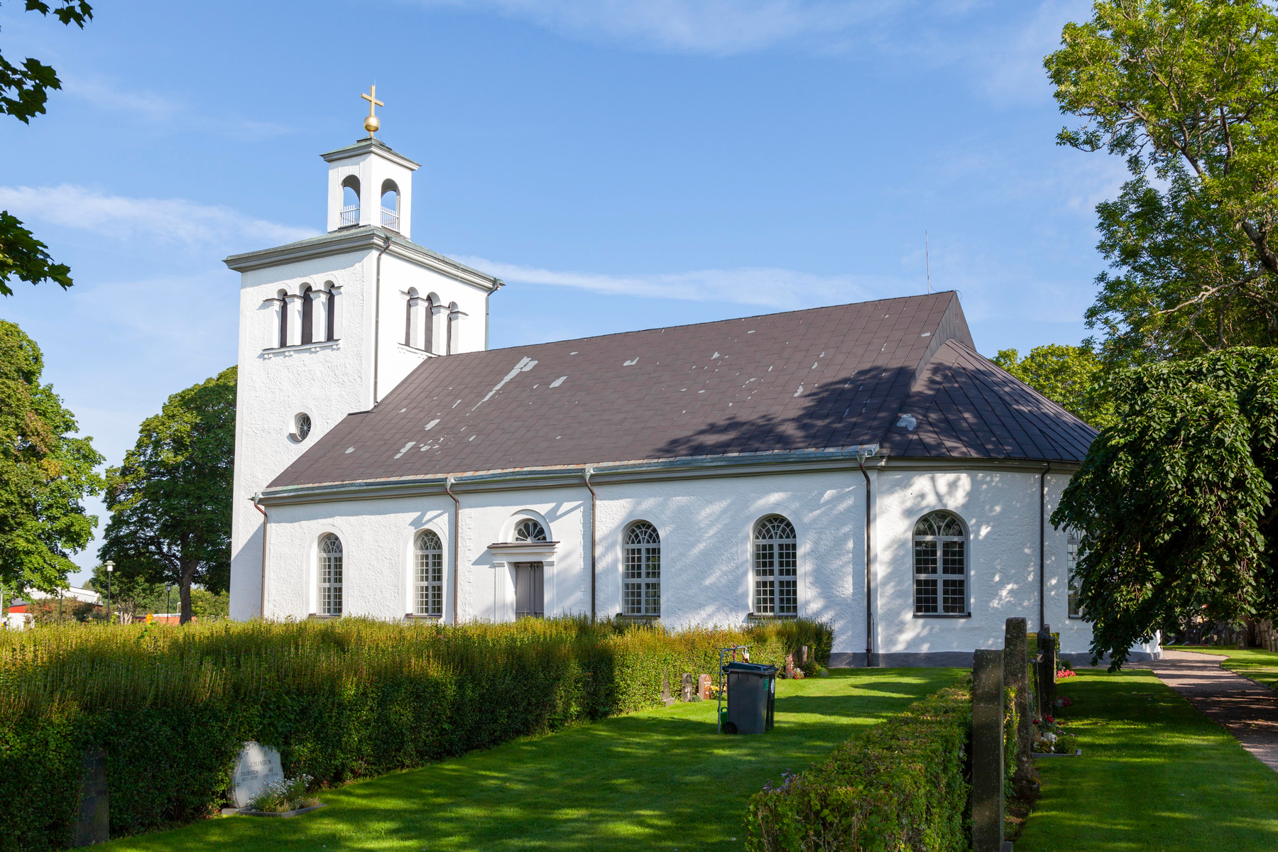 En vit kyrka med ett brunt tak och en vit kyrktorn med ett gyllene kors på toppen, omgiven av gröna träd och en gräsmattapå en solig dag.