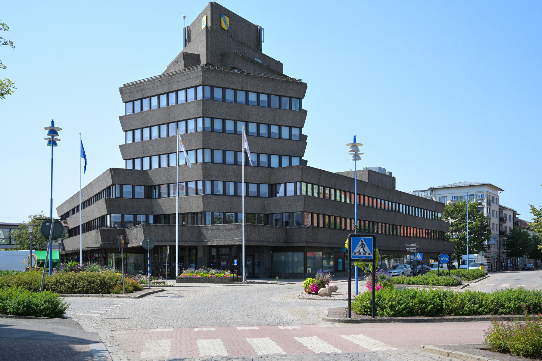 En modern byggnad med en komplex arkitektur. Byggnaden är grå och har flera våningar med många fönster. Utanför byggnaden finns flaggstänger med flaggor, buskar, blomarrangemang och en trottoar med övergångsställe. Himlen är klarblå.