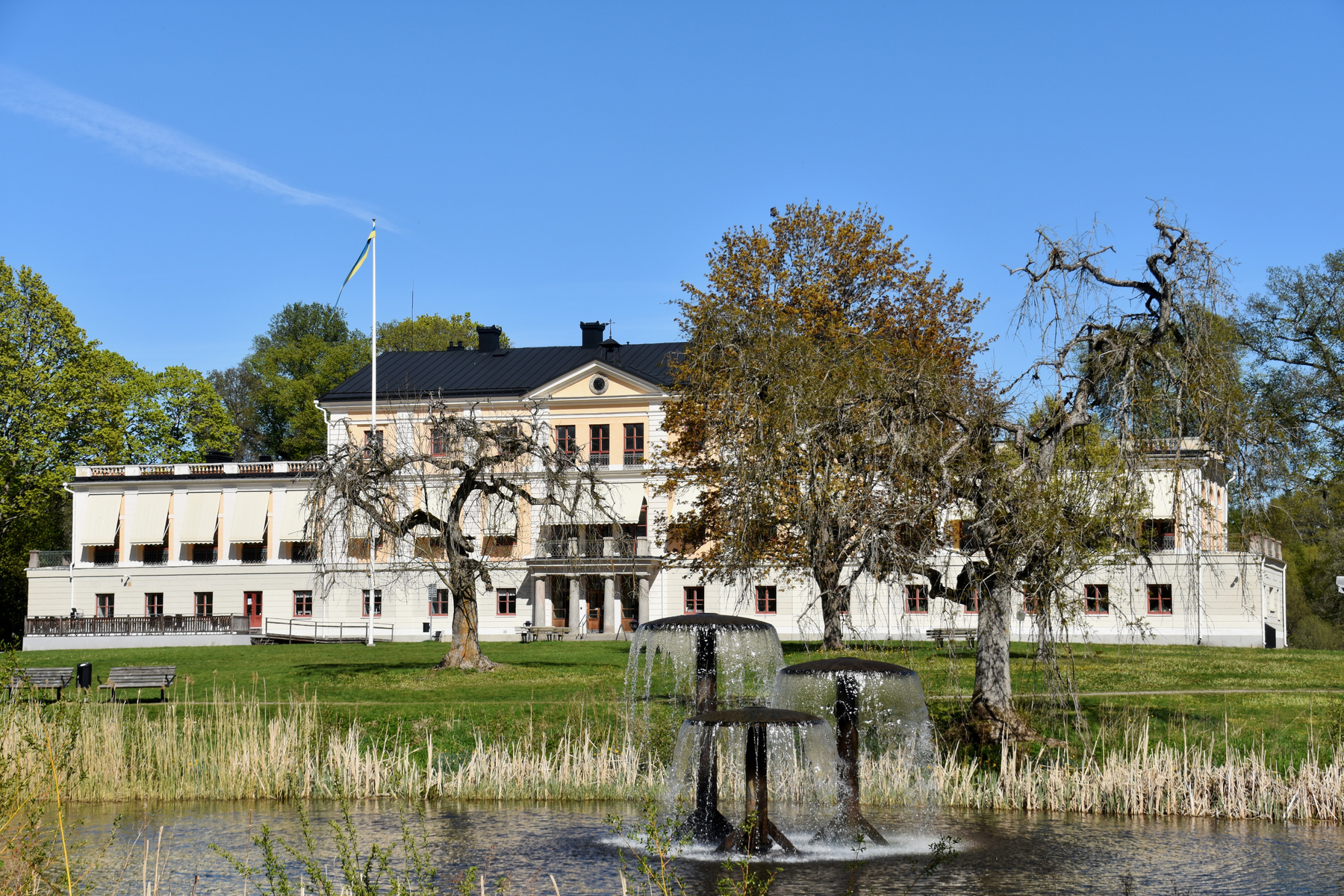 En stor, ljus byggnad med mörk tak och röda fönsterkarmar står bakom en gräsmatta. Framför byggnaden finns flera träd, varav några är kala. I förgrunden syns en damm med tre fontäner och vass längs kanten. En svensk flagga syns vid byggnadens vänstra
