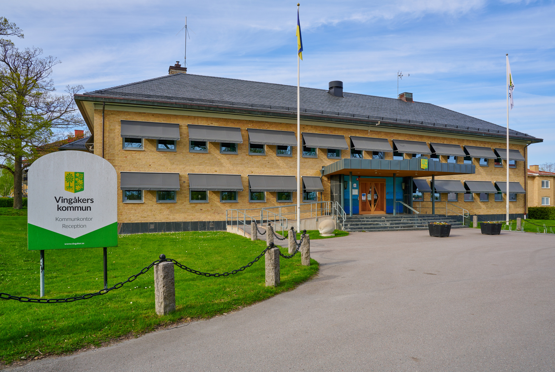 En gul tegelbyggnad med grått tak och grå fönstermarkiser under en blå himmel med några moln. Framför byggnaden finns en gräsmatta, en asfalterad gångväg, samt en skylt som anger "Vingåkers kommun". En svensk flagga syns också på en flaggstång.