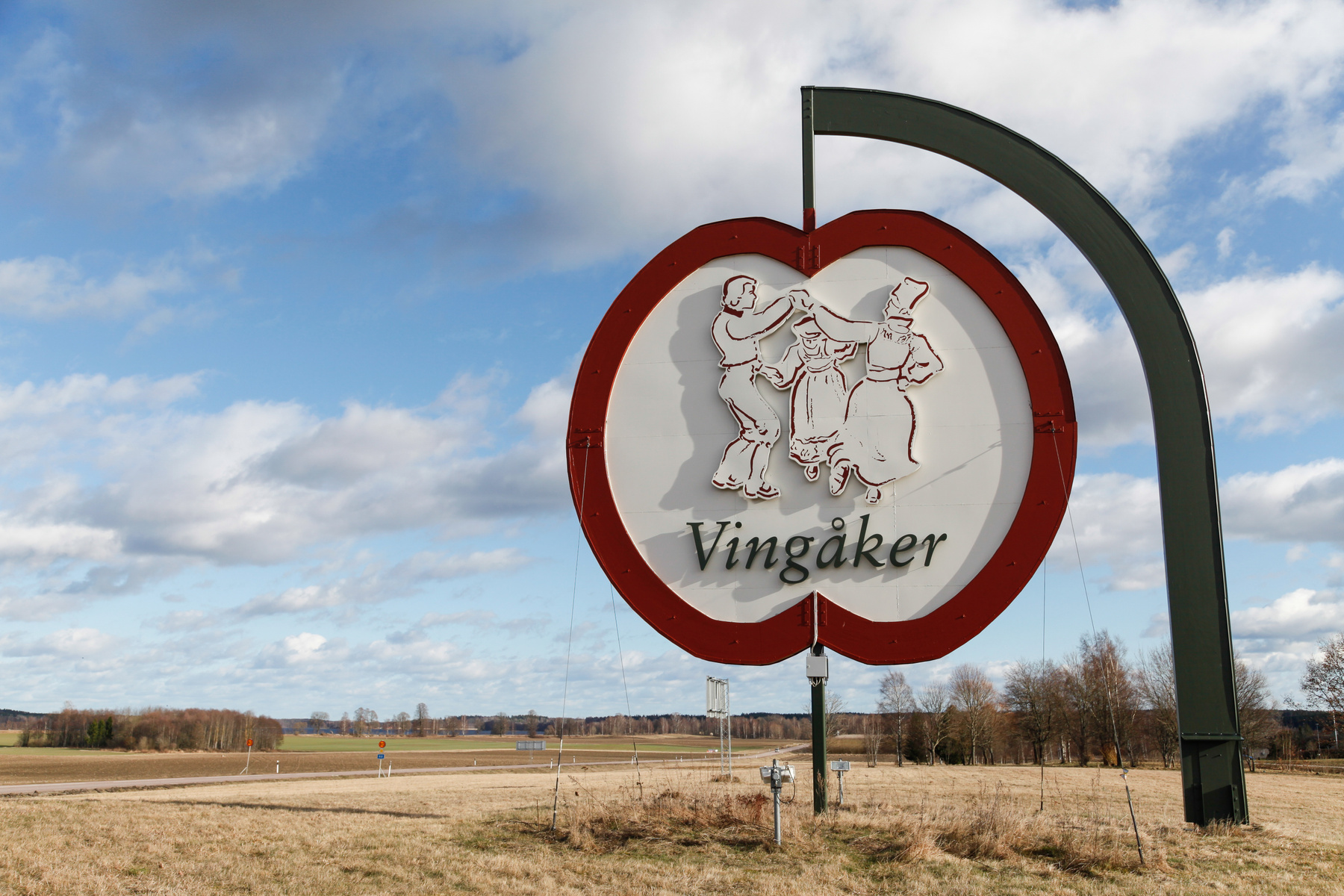 En stor äppelformad skylt med texten ”Vingåker” och ett motiv av tre folkdansare står i förgrunden. I bakgrunden syns ett öppet landskap med åkrar, en väg och träd under en delvis molnig himmel.