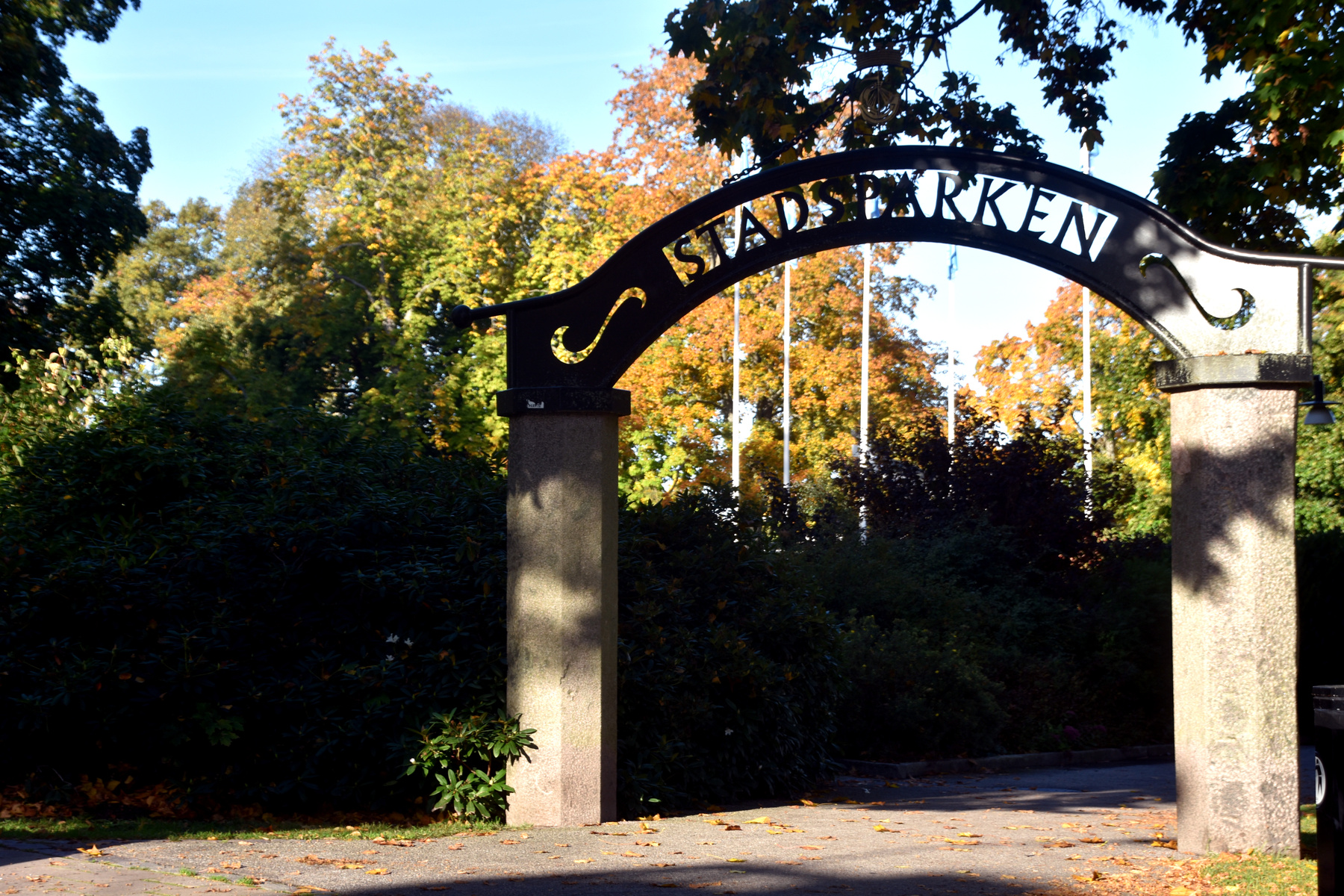 En bild tagen utomhus under dagtid, som visar en parkentré med en svart smidesportal där ordet "STADSPARKEN" står skrivet i versaler. Portalen vilar på två stenpelare. I bakgrunden syns träd med höstlöv, en del i gult och orange. Framför portalen fin