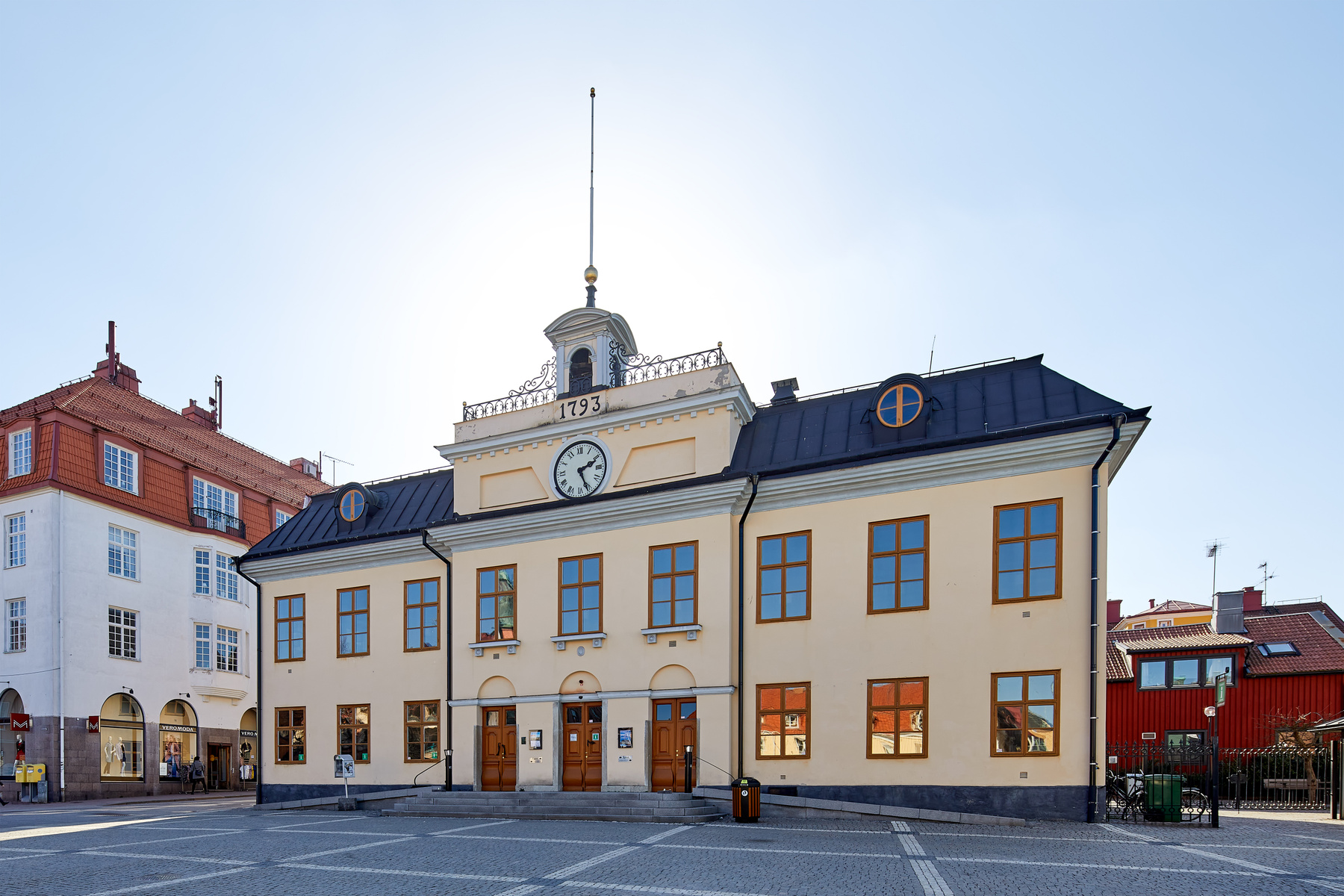 En gul byggnad med klocka och "1793" ovanför huvudentrén under en klarblå himmel. Till vänster syns delar av en vit byggnad med rött tak och butiker i markplan. Framför byggnaderna ligger ett torg belagt med gatsten. Solen lyser från höger. Flera fön