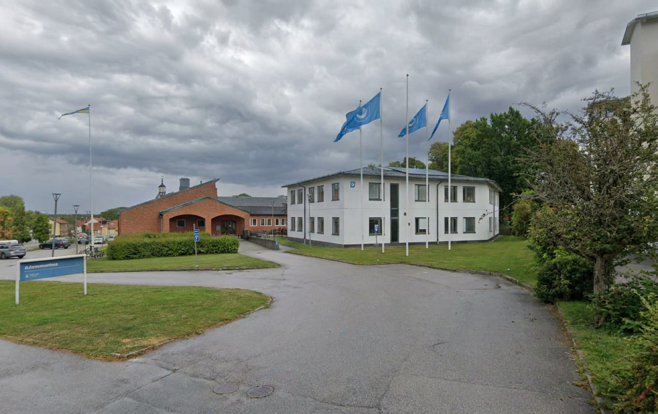 En utomhusbild som visar en parkeringsplats med flera byggnader och grönområden. Till vänster finns en röd tegelbyggnad och till höger en vit byggnad med flera flaggstänger framför. En blå kommunskylt med texten "Kommunkontoret" syns i förgrunden.