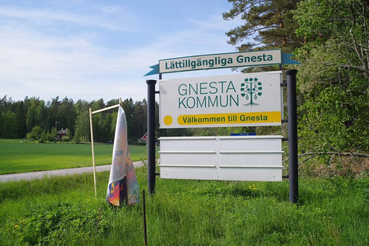 En vägskylt med texten "Lättillgängliga Gnesta" och "GNESTA KOMMUN Välkommen till Gnesta" står vid sidan av en väg. I bakgrunden syns ett grönt fält, träd och ett blått himmel med vita moln. En mindre färgglad flagga sitter bredvid skylten.