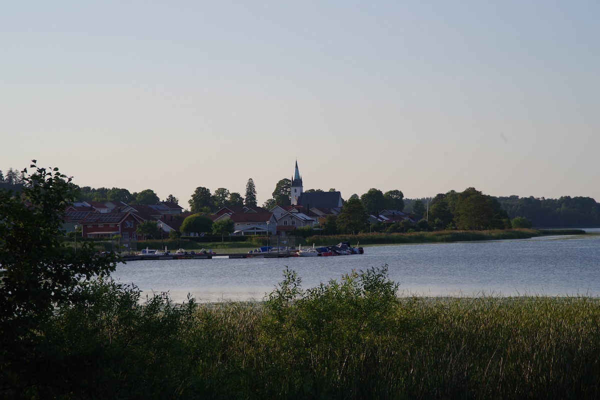 En översiktsbild av en sjö med ett samhälle längs strandlinjen i bakgrunden. Ett antal hus med röda tak syns, och i mitten av samhället reser sig en kyrka med en spetsig spir. Längs sjökanten ligger en brygga med båtar förtöjda. I förgrunden syns grä