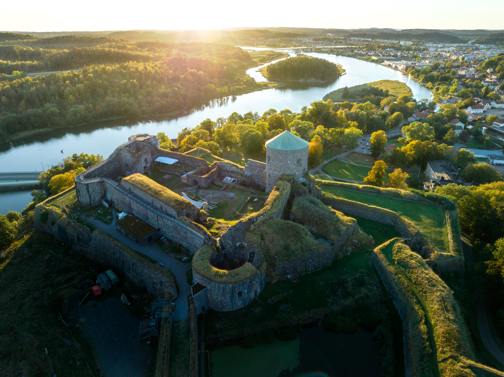 En flygfotografisk vy av en gammal fästning med stenväggar och gräsbevuxna takbeläggningar. Framför fästningen slingrar sig en flod genom ett landskap med skog och bebyggelse, allt under en solnedgång eller soluppgång som skapar ett gyllene ljus.