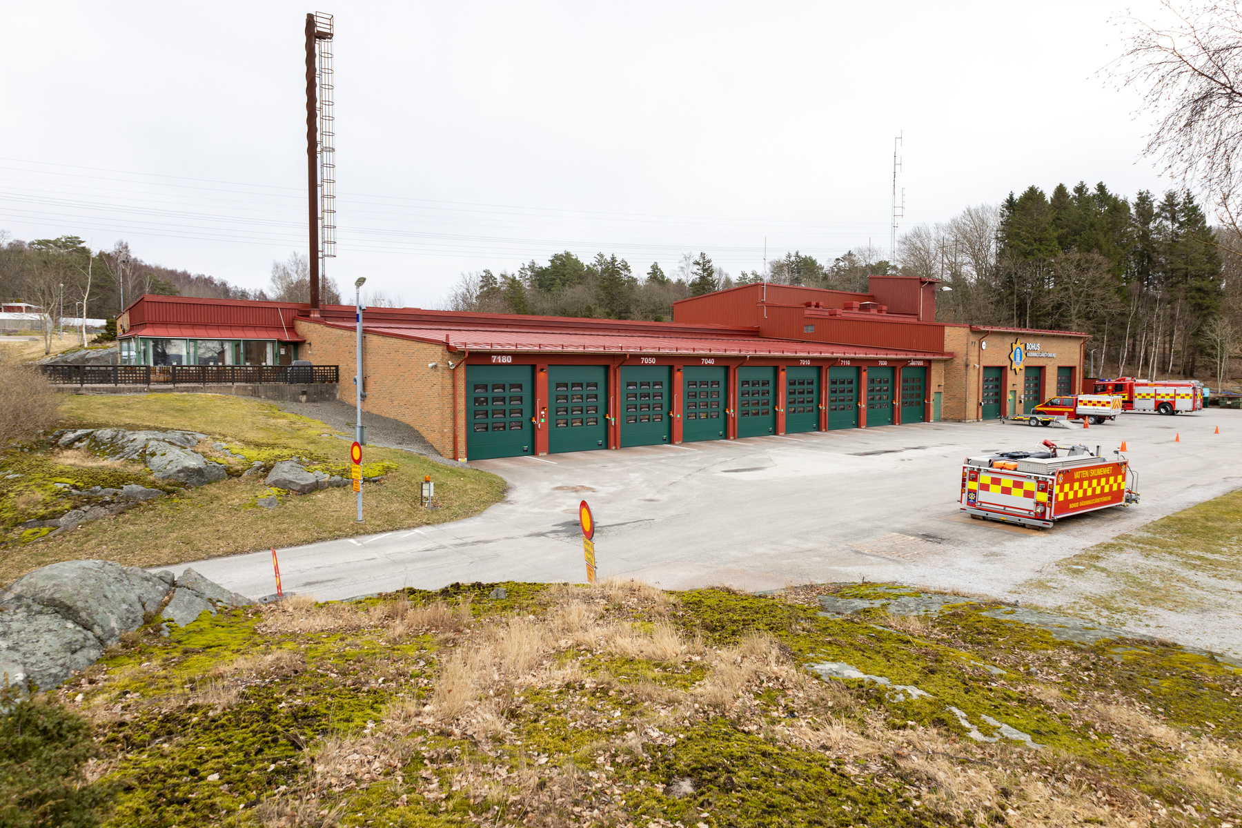 En fasadvy av en brandstation med röda tak och tegelfasader. Flera gröna garageportar är synliga, varav flera har nummer skrivna ovanför. På gården står en räddningsbil och en personbil med släpvagn, samt en mindre "Vatten Skutan"-enhet. Området omge